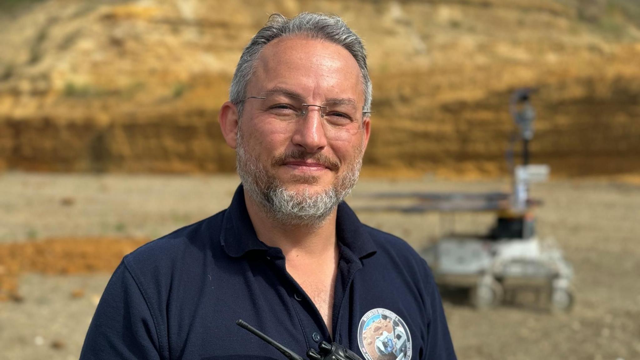 A man wearing an Airbus branded navy polo shirt is smiling at the camera. He has grey hair and a grey beard. A rover is behind him in the quarry he is standing in.