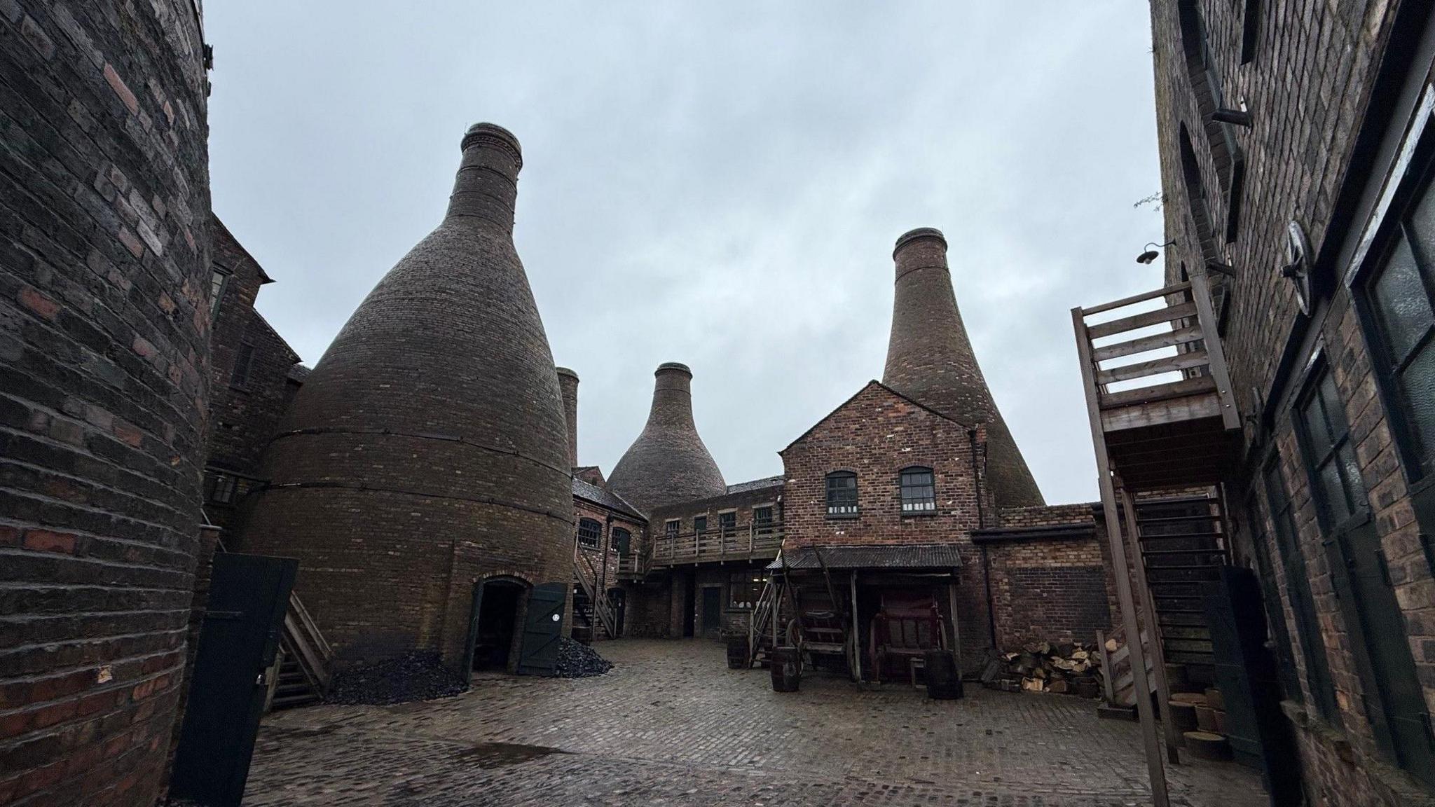 A cobbled courtyard surrounded by brick-built buildings and historical bottle ovens. There are a number of outbuildings and piles of coal located alongside the bottle ovens.