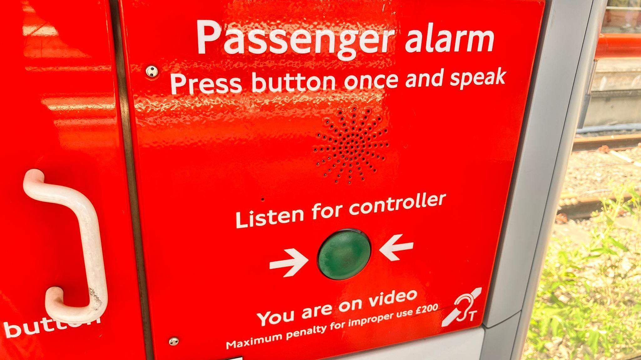 A red emergency panel with a green button in the center, labeled for passenger use. Instructions warn that misuse carries a £200 fine and that the user is on video.
