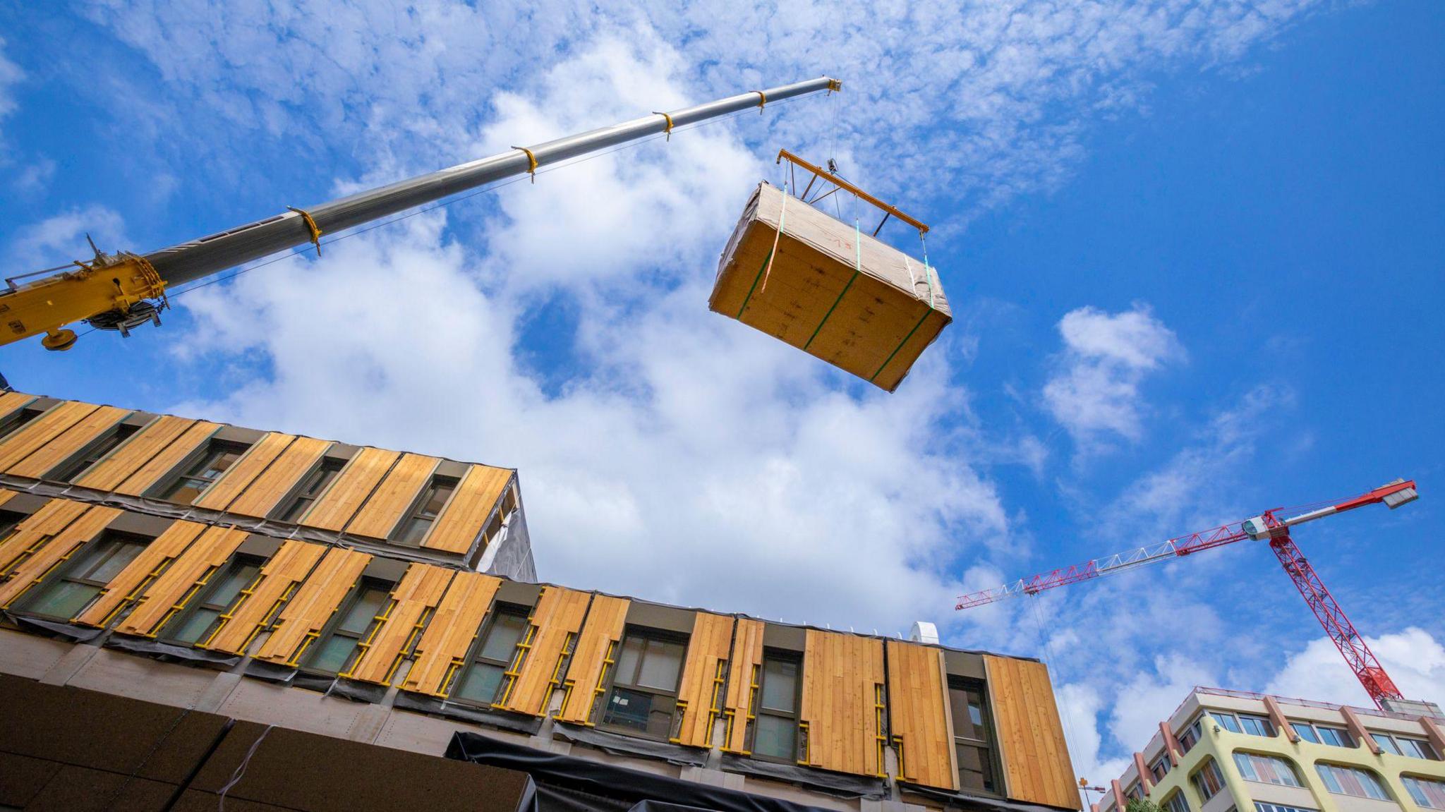 A crane is lifting a large wooden box at the side of a modular building.