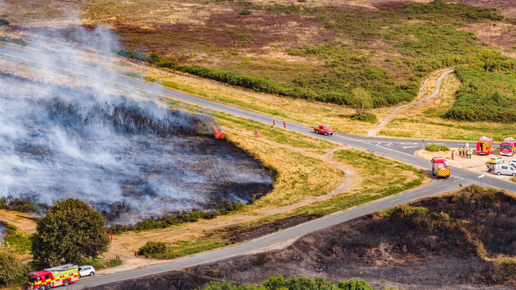 Fire engines parked on roads around a heath which is black from fire, pockets of flames can be seen and large amounts of smoke.