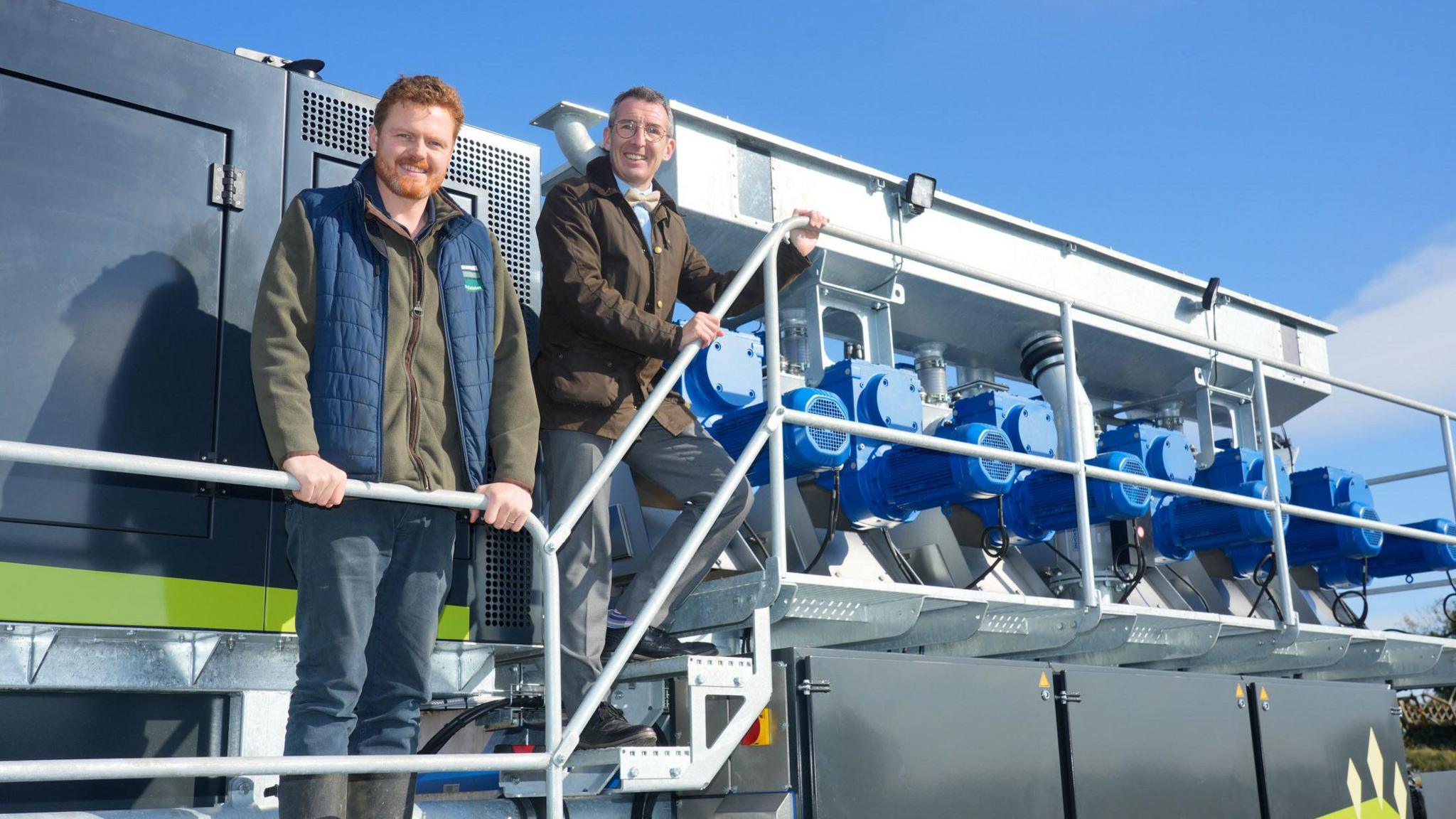 Andrew Muir with Jack Blakiston Houston from BH Estates. Andrew Muir is wearing a brown coat, light blue shirt, yellow bow tie and glasses. Jack Blakiston Houston has ginger hair and beard, wearing a blue puffer vest and green jumper. Both are standing on the steps of farm equipment holding the railings.