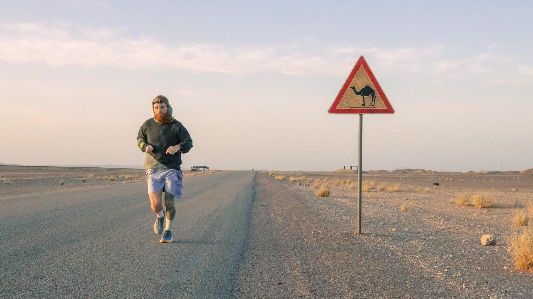 A man running along an empty road, with a sign to his side. He is wearing a grey hooded top and blue shorts. 