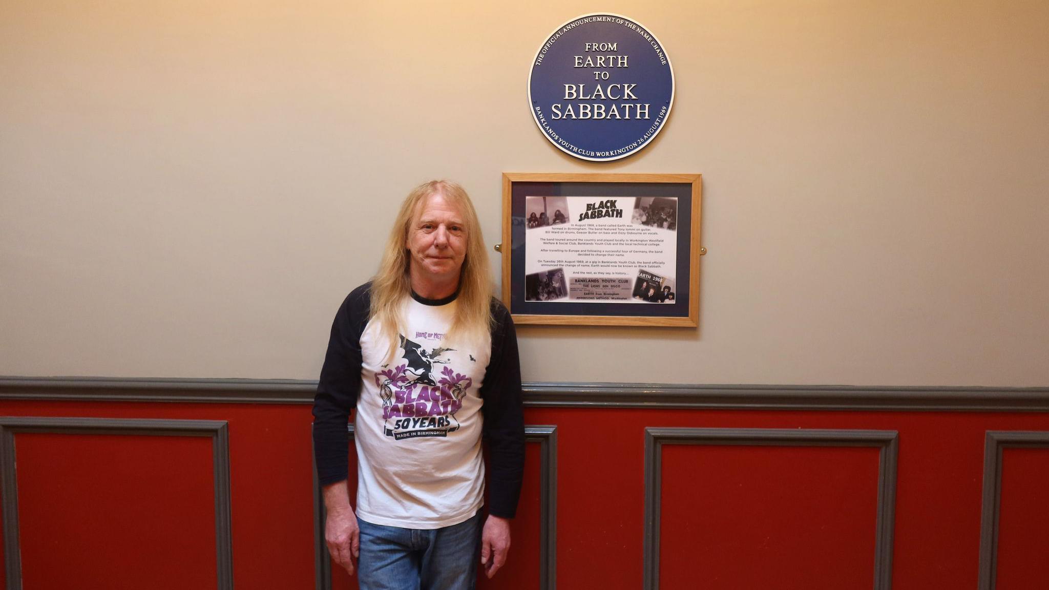 Des Rumney with blonde hair, wearing a Black Sabbath long-sleeved top and jeans, stands next to a blue plaque which reads 'Black Sabbath'.