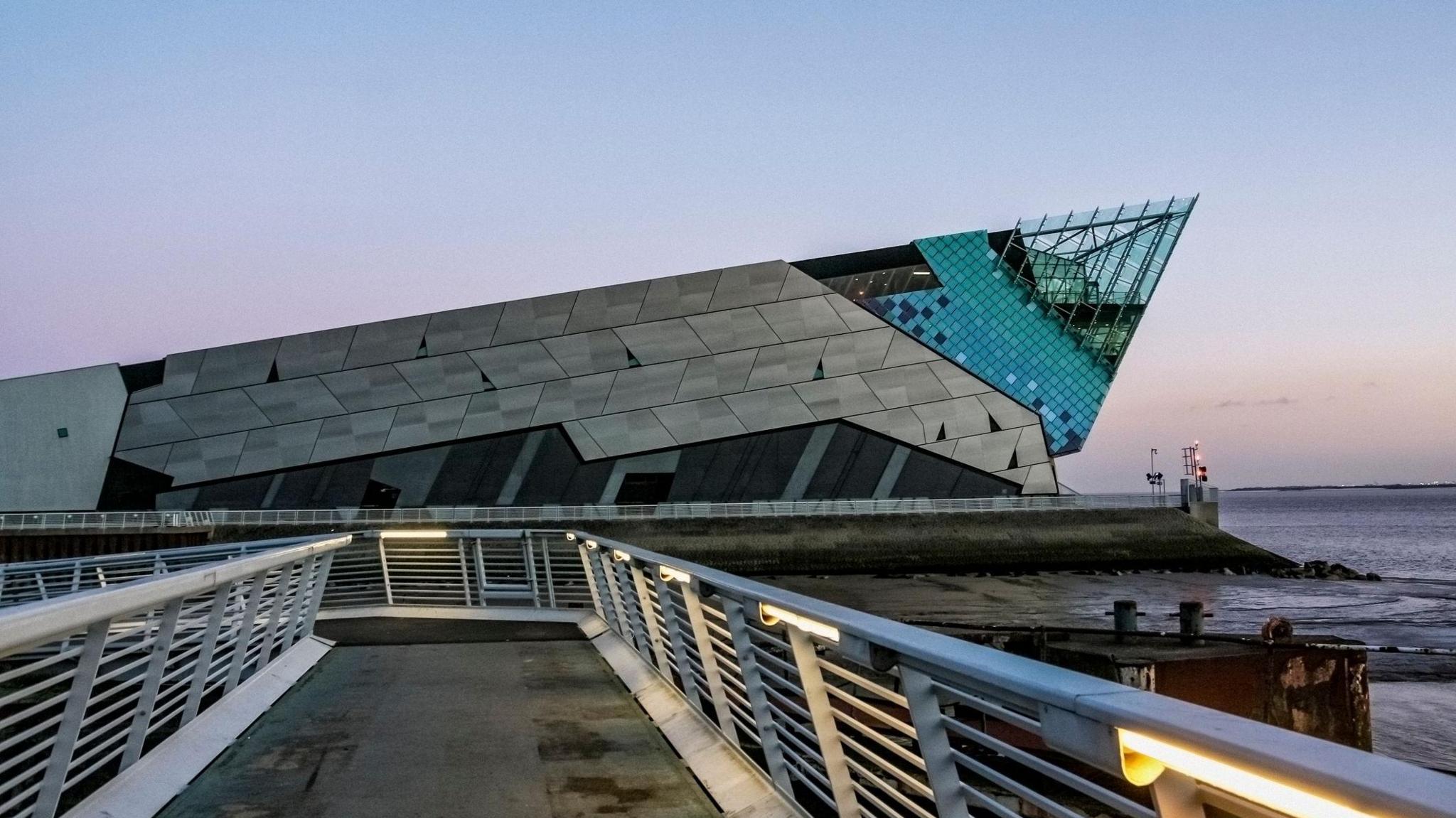Building clad in metal panels is shown facing the water, with a glass section pointed towards the sky. Leading up to the building is a metal walkway with railings on both sides. 