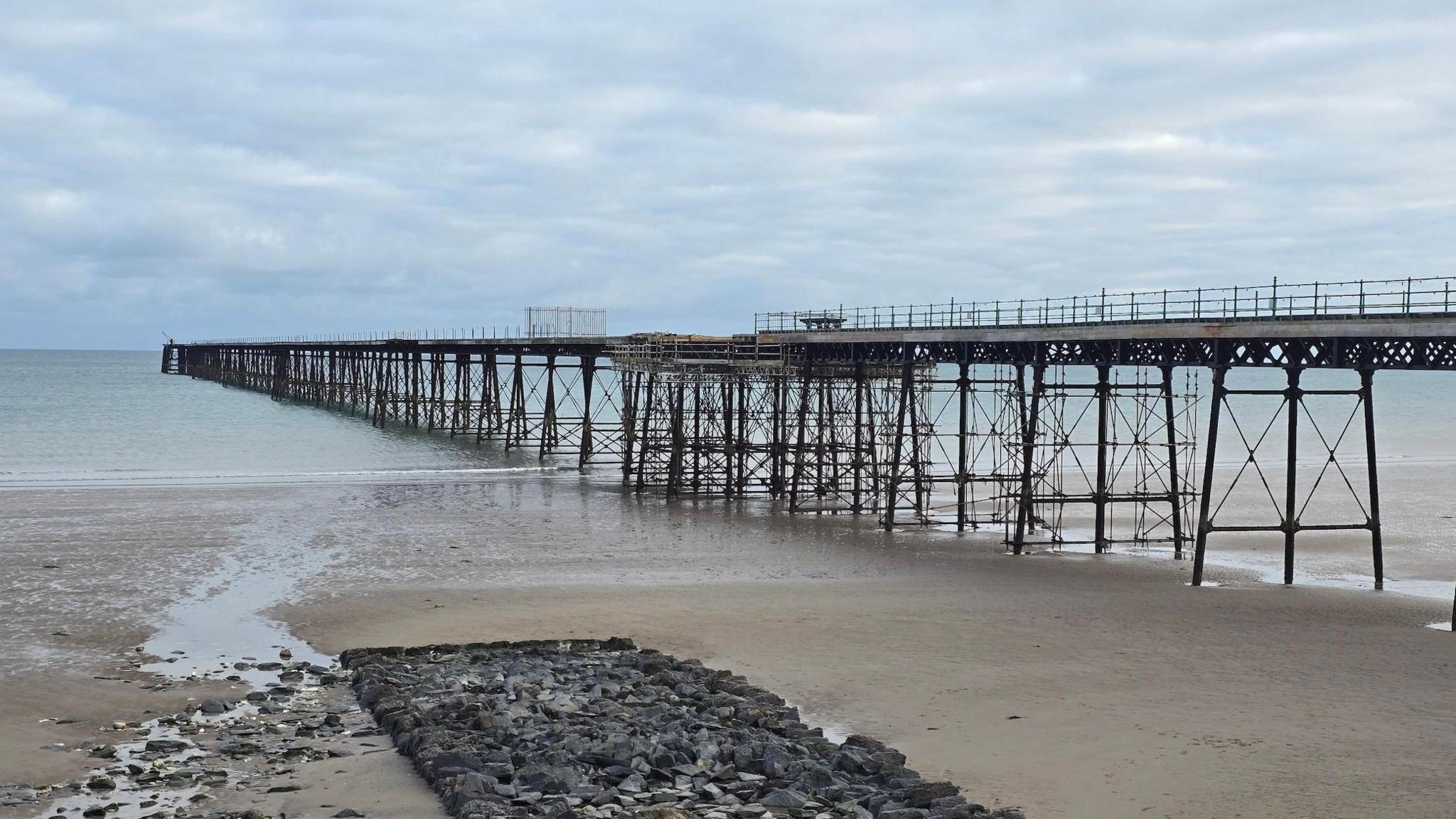 The iron pier runs from the right to the left down a sandy beach into the sea in the distance. There is a large cluster of rocks on the sand to the left. It is a cloudy but fairly bright day and the sea and very calm.