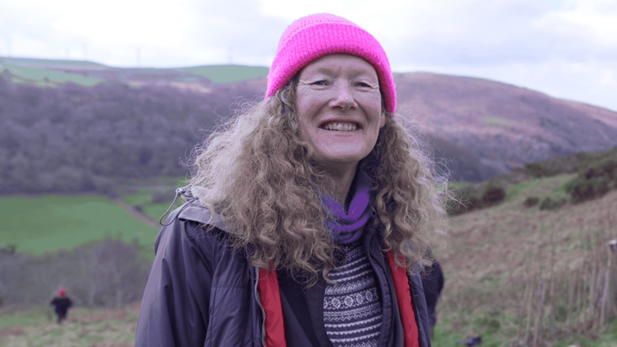 Llun o Linda Denton - cydlynydd y gwirfoddolwyr. Mae'n gwisgo het binc ac mae ganddi wallt hir cyrliog, ac mae'n sefyll ar ochr bryn ger Tal-y-bont yng Ngheredigion 