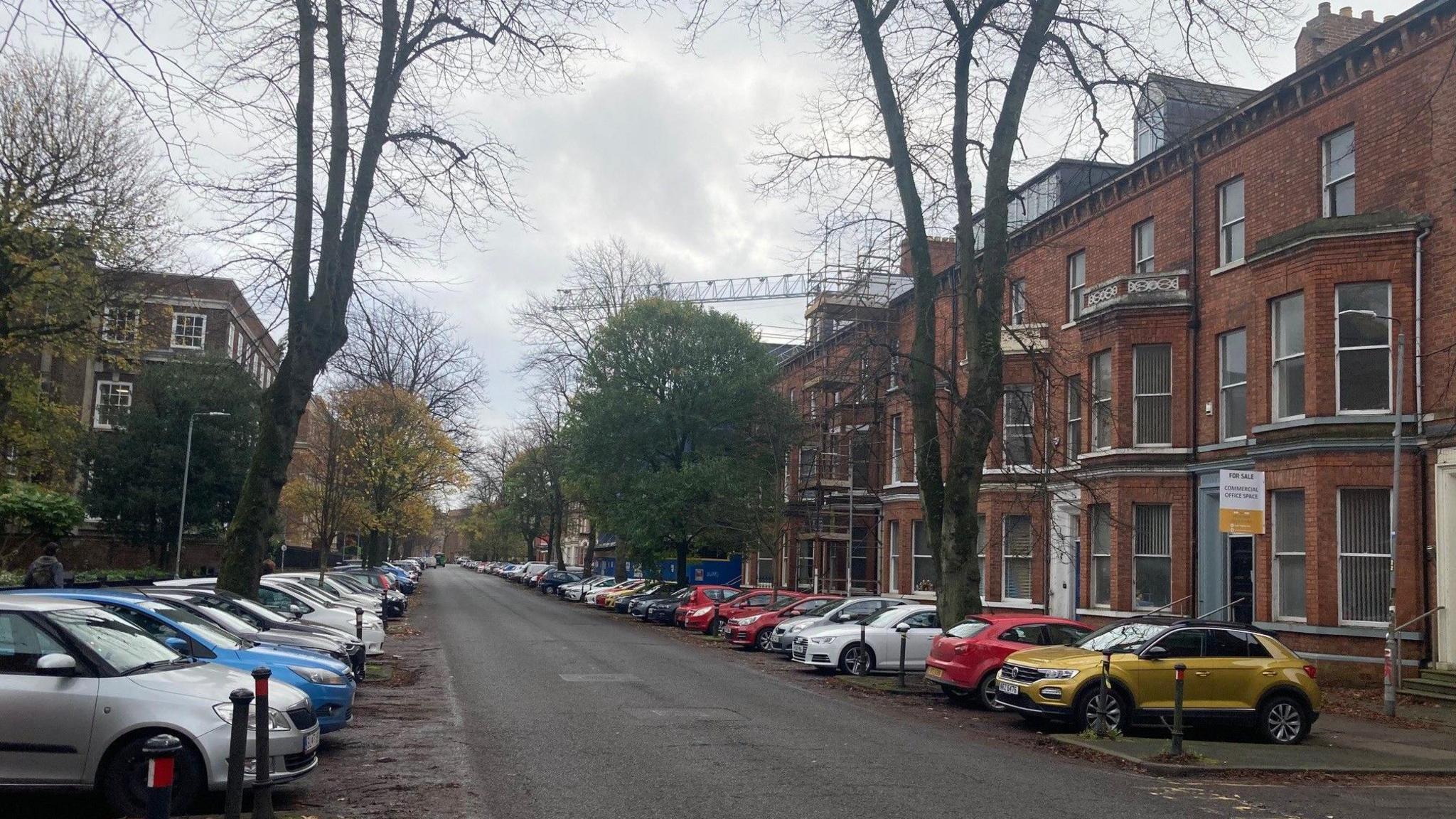 An empty road with cars parked in spaces on either side. On the right side of the road, there is a row of re-brick buildings, some of which have scaffolding. On the other side there are larger buildings. There are trees on both sides.  