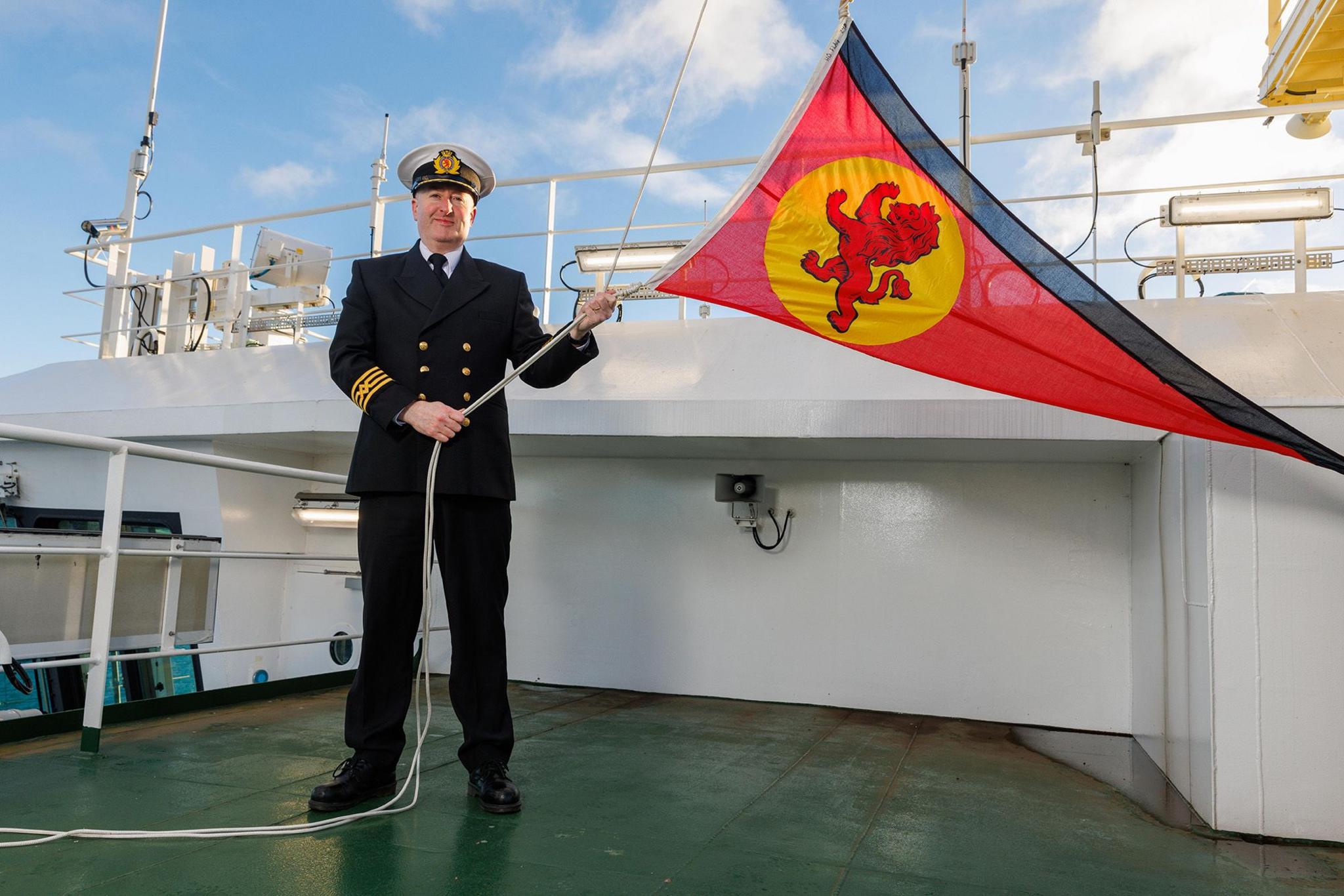 A man in naval uniform raises a red ensign with a rampant lion motif on the deck of Glen Sannox