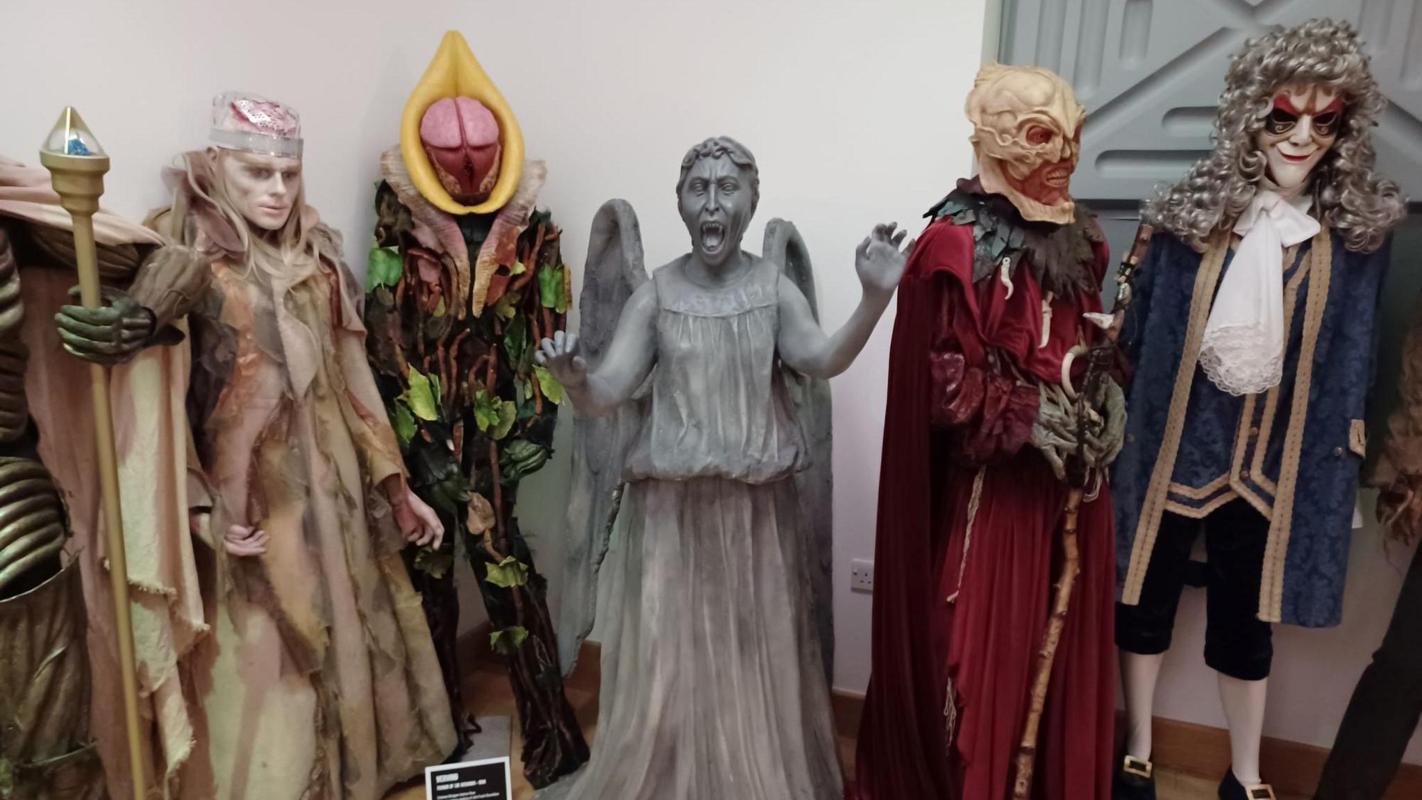 An exhibition display of mannequins dressed to appear as various creatures from the BBC science fiction series Doctor Who. The central figure and focus of the photo is a Weeping Angel, a creature designed to look like a stone statue of the type which might be seen in a graveyard. It has its arms raised and spread, and its mouth open to reveal a sharp-toothed scream.