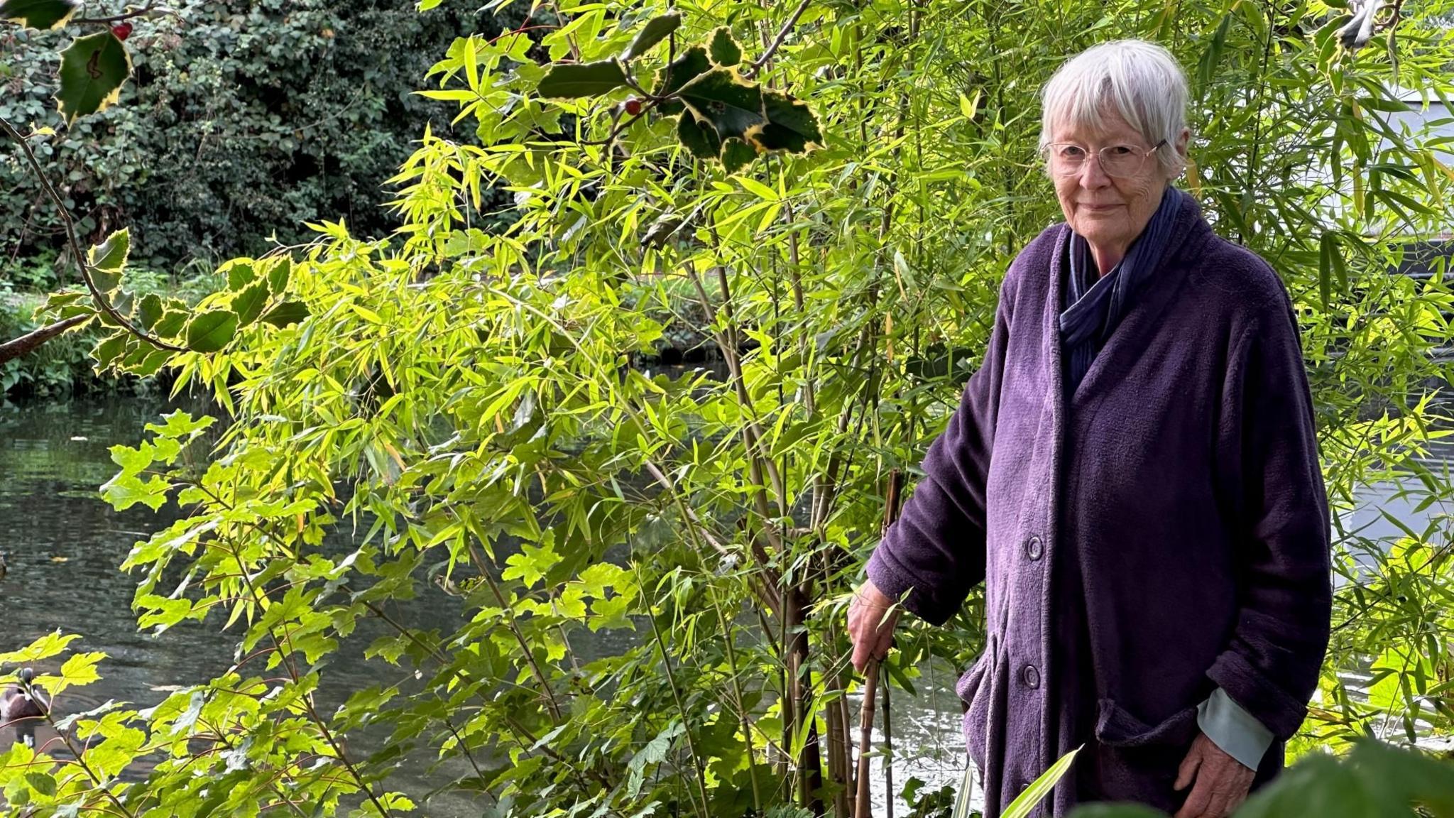 A grey-haired woman in a purple coat stands on the edge of the canal from her garden.