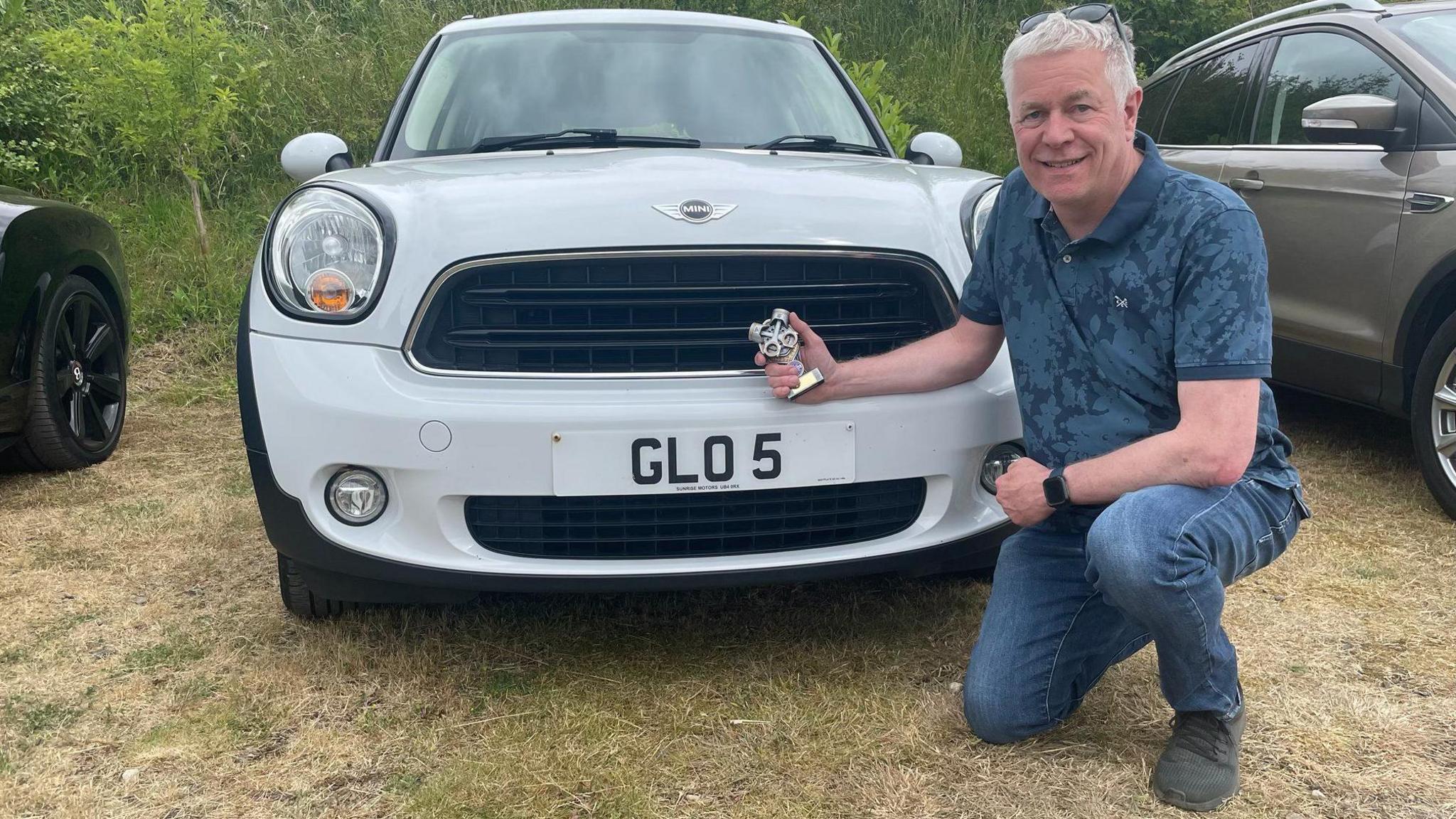 Neal Bircher poses on bent knee in front of his white Mini that has a 'GLO 5' number plate