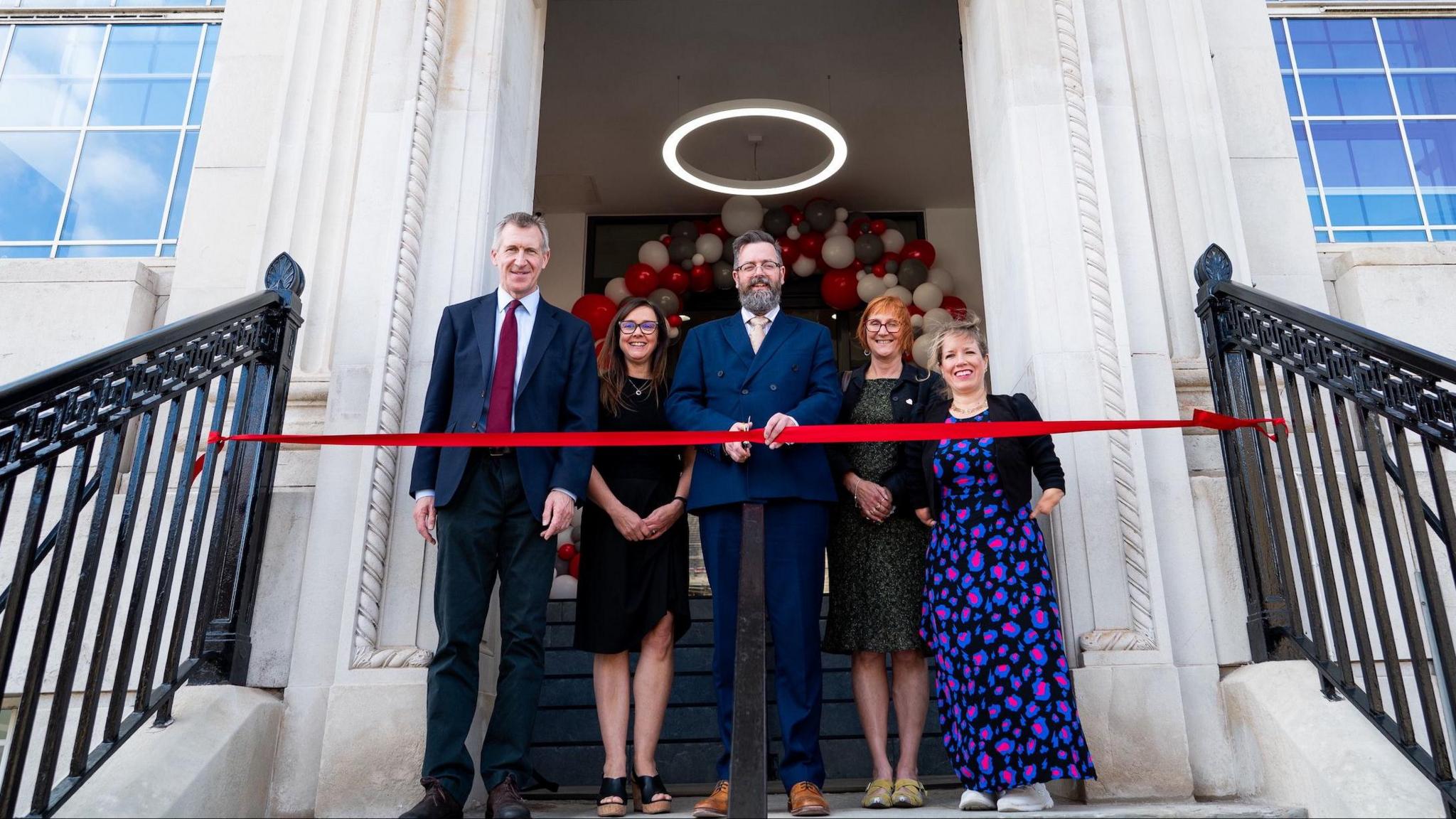 A group of smartly dressed adults stand infront of a red ribbon which is stretched out infront of a fine white stone building