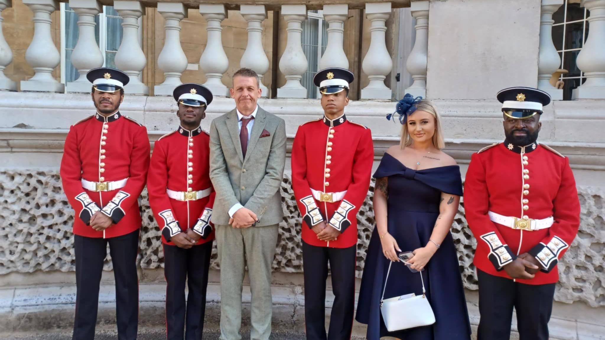 Dave wearing a beige suit, white shirt and a red tie and pocket square, standing with four soldiers in red coats, and his daughter who is in a blue dress wearing a blue fascinator hat.