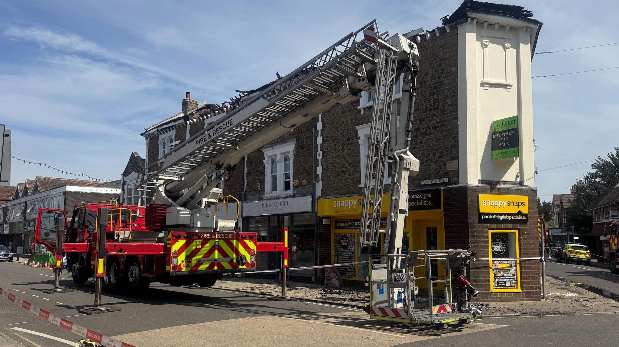 Fire appliance with aerial ladder parked outside a terrace of shops with flats above with its roof missing.