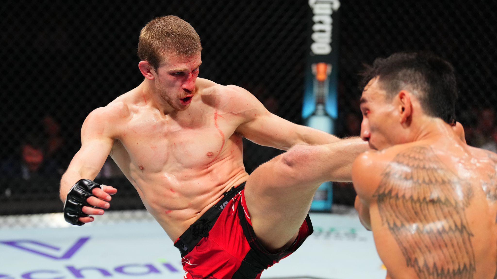 Arnold Allen is pictured in an MMA fighting ring during a match against a tattooed fighter. Allen is wearing red and black shorts as well as black gloves. He has a bloody face and there are also traces of blood on his chest. 