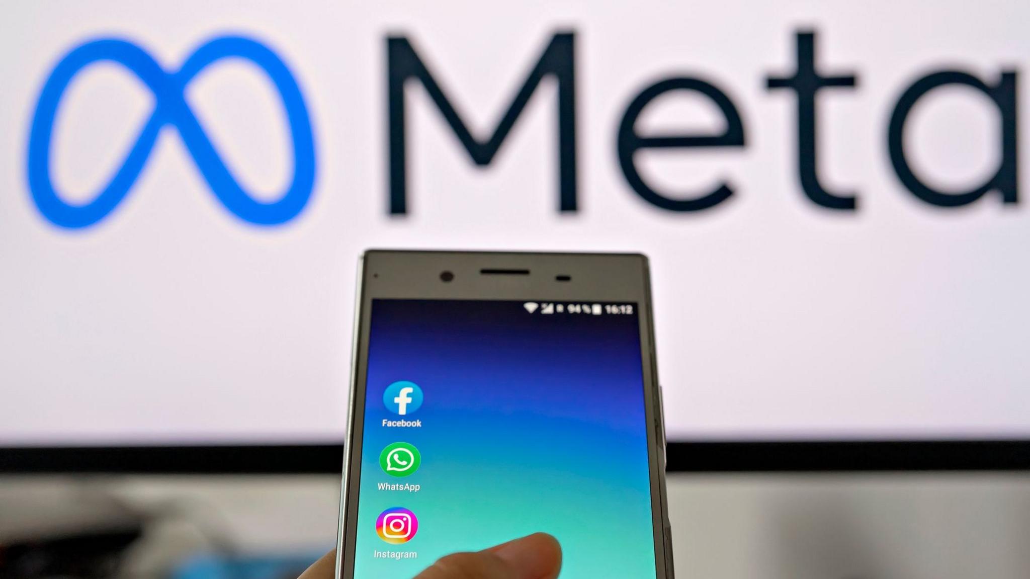 A white screen in the background shows the Meta logo and in front of it someone is holding a phone with the Instagram, Facebook and Whatsapp apps on it.