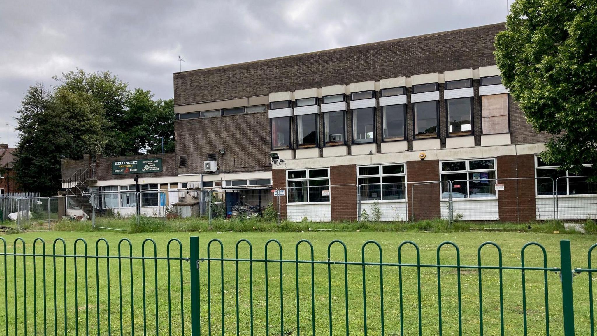 A brutalist building pictured from behind a green fence. Some metal fencing stands closer to the premises.