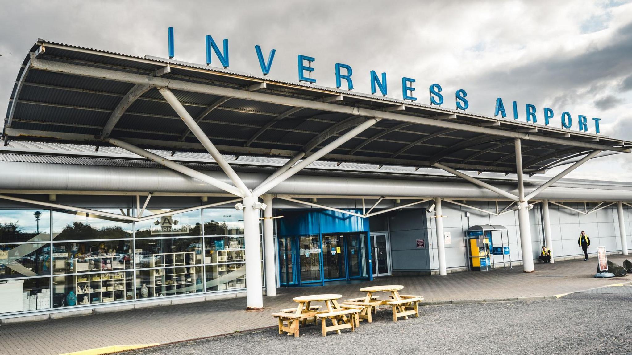 The main entrance to Inverness Airport. The name of the airport is spelled out in large lettering on a metal canopy above a blue revolving door.