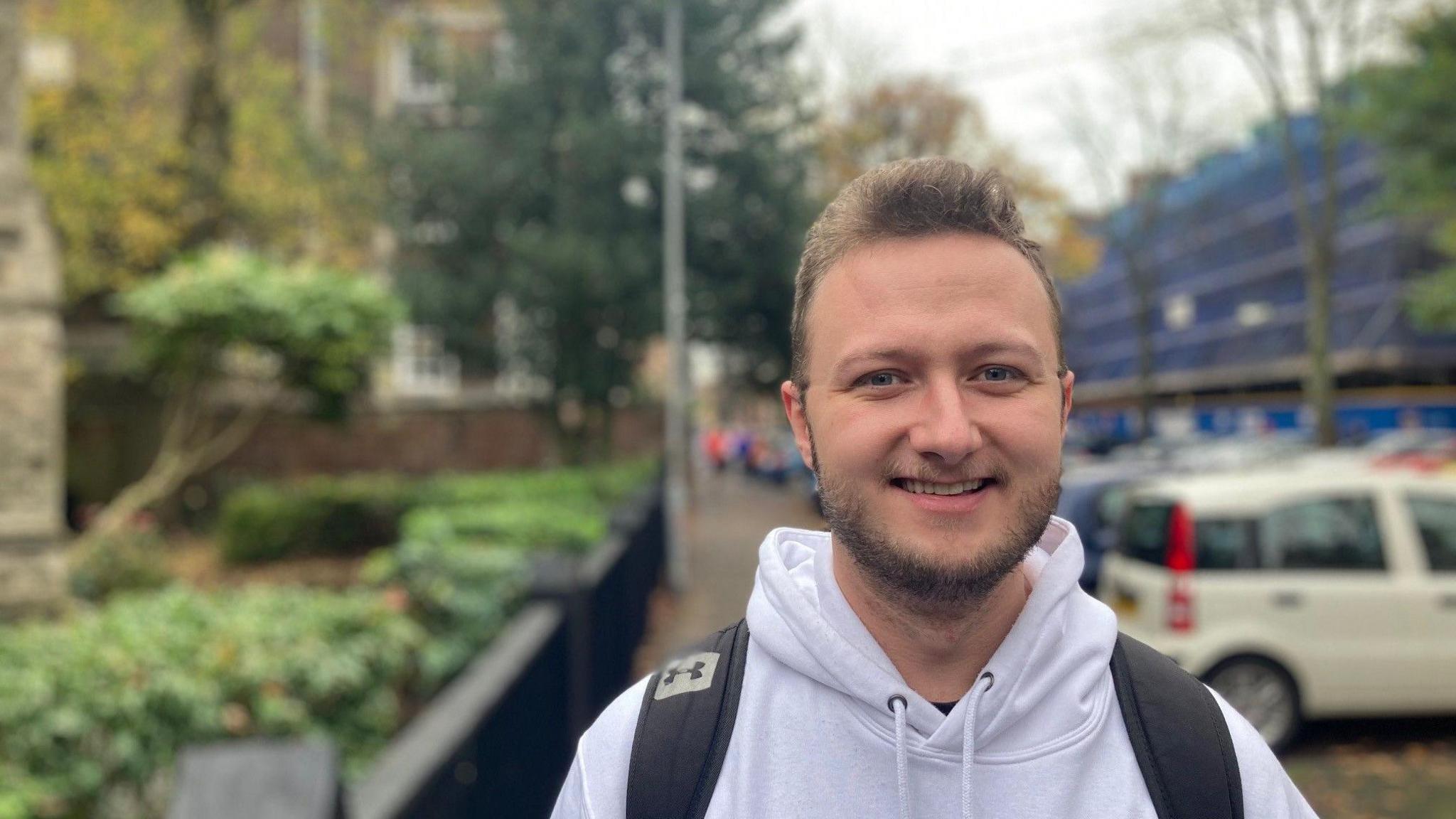 A man in a grey hoodie smiles at the camera. He has short brown hair. He is wearing a backpack, and the black straps of the backpack are visible over his shoulders. In the background is a row of parked cars, some trees, some buildings, and some blue scaffolding. 