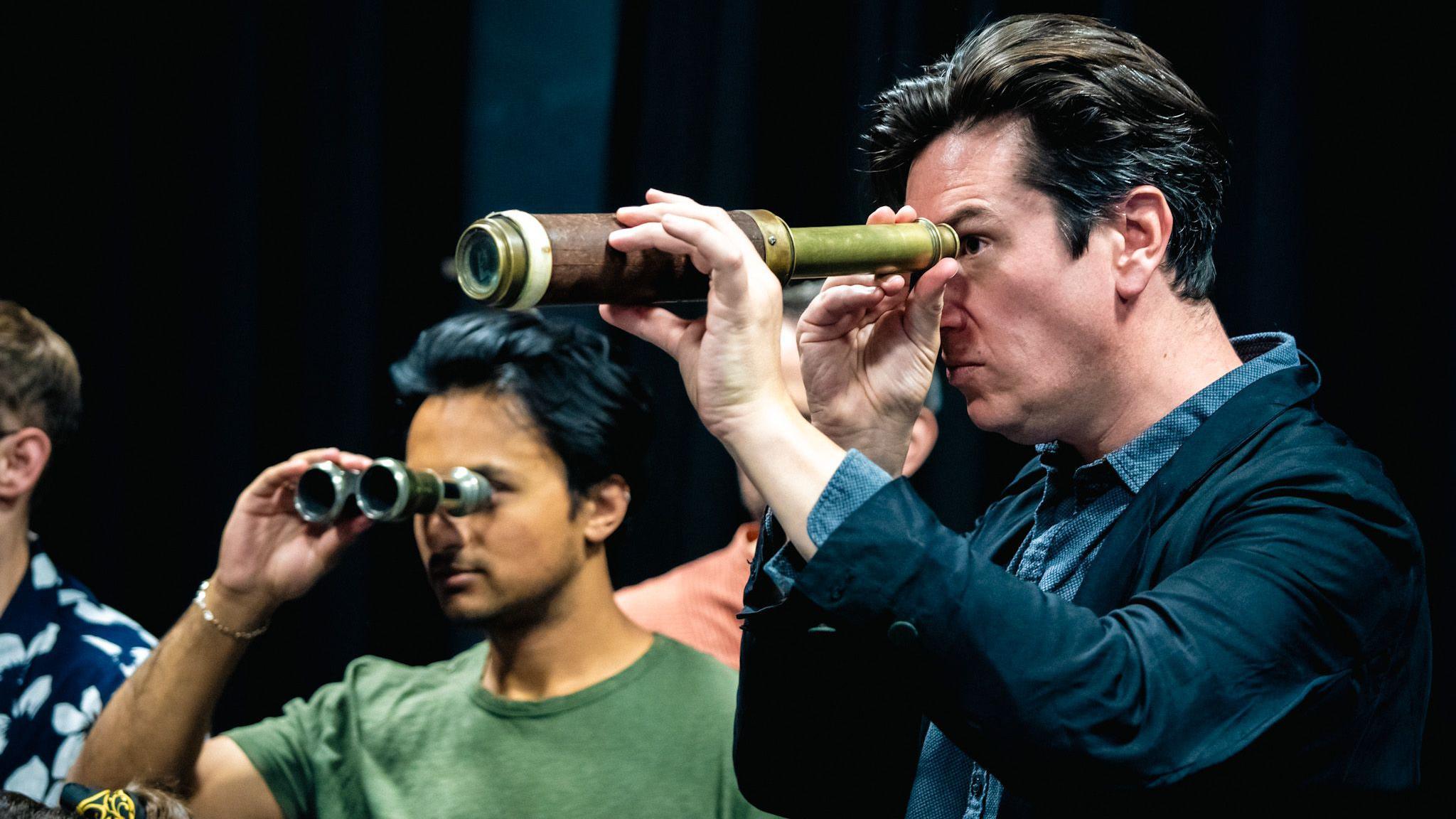 Ed Hawkins looking through a telescope during a rehearsal. He is wearing a blue shirt and a dark blazer. Other cast members can be seen in the background, including one looking through a pair of binoculars