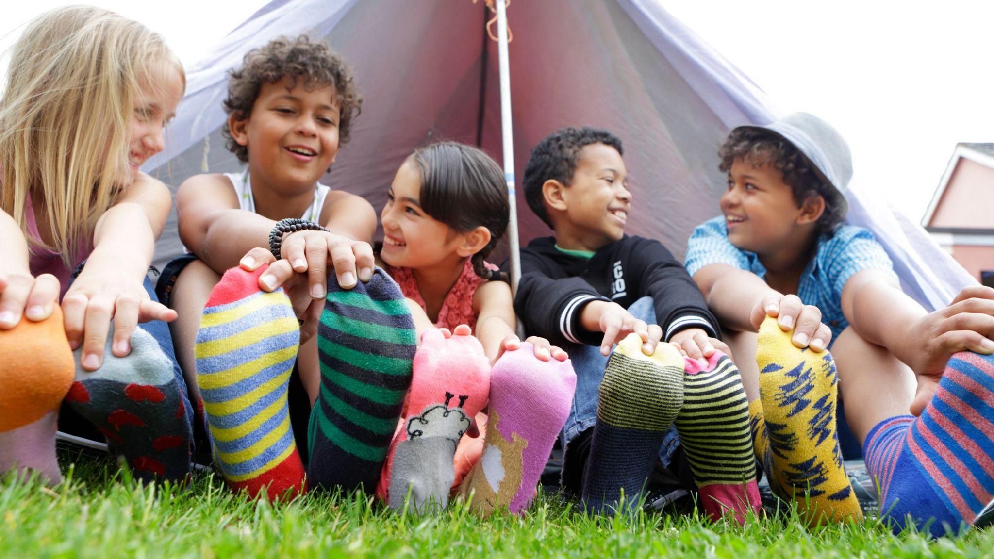 Children are sitting outside a tent on the grass. Their legs are outstretched and they are all wearing brightly coloured odd socks.