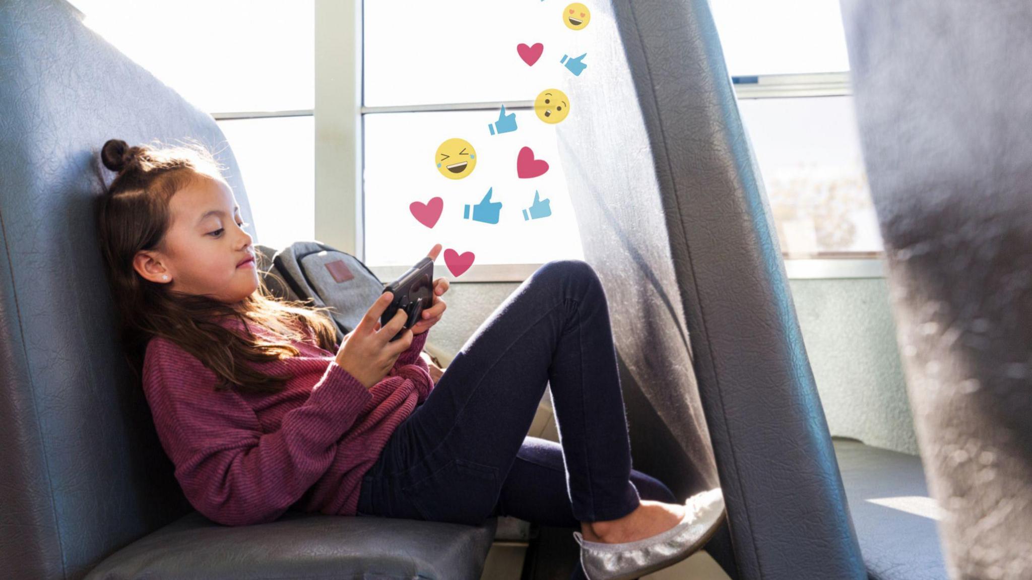 Young schoolgirl relaxes on the school bus. Graphics of emojis, like buttons and heart symbols flow out from her smartphone.
