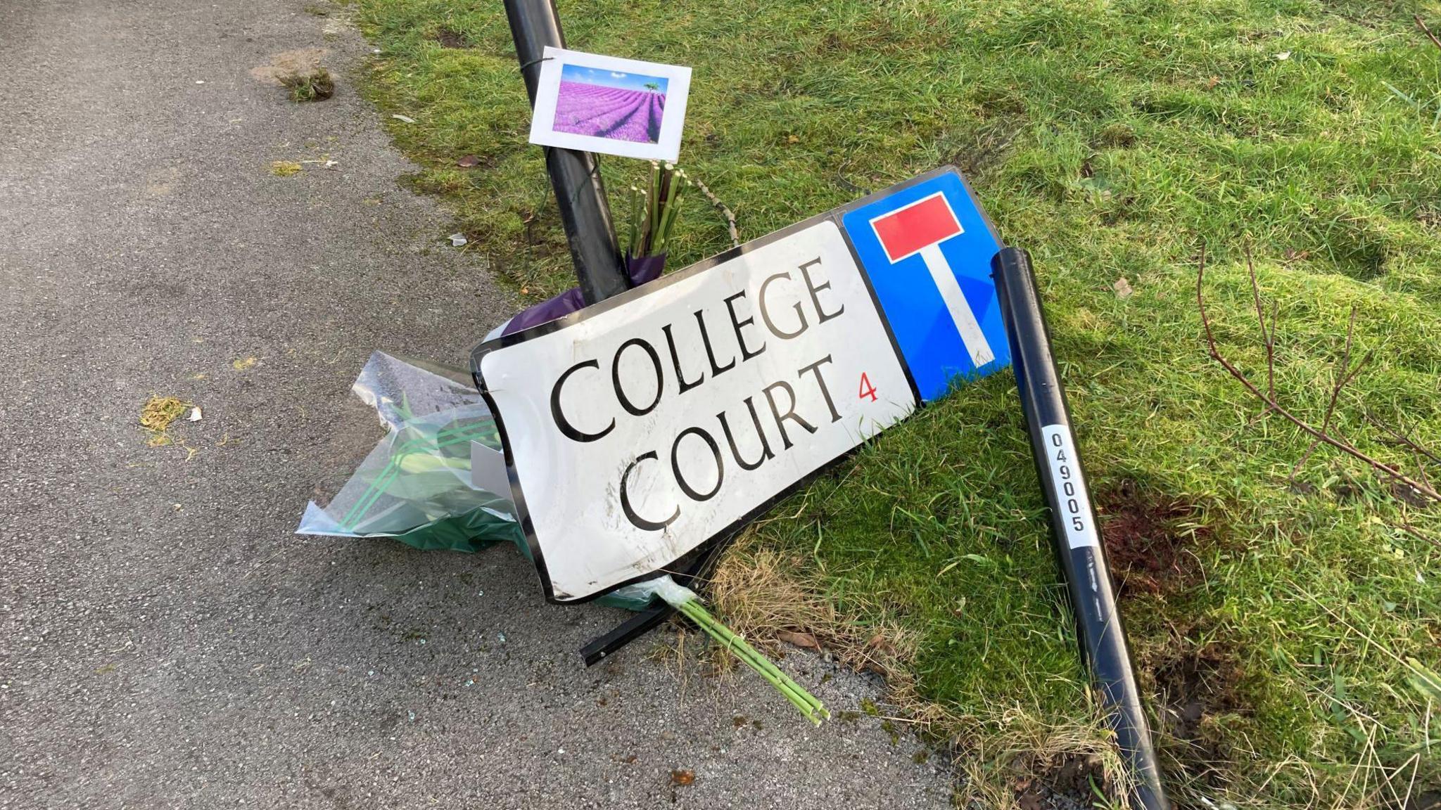 Flowers are left next to a road sign displaying "College Court", which has been knocked over.