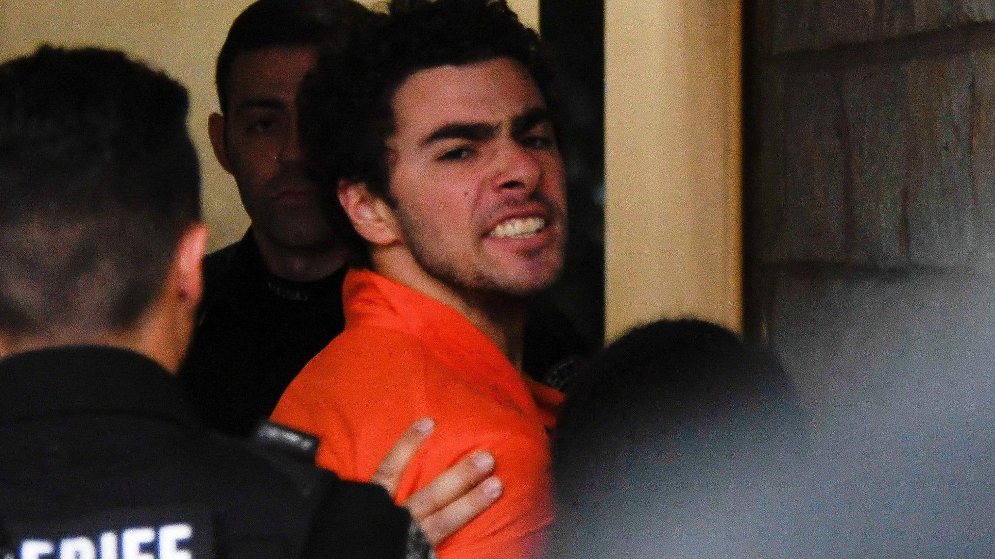 Luigi Mangione, 26, a suspect in the New York City killing of UnitedHealth executive Brian Thompson, is seen looking angry with deputies in an orange prison jumpsuit. He is at court in a hallway.  