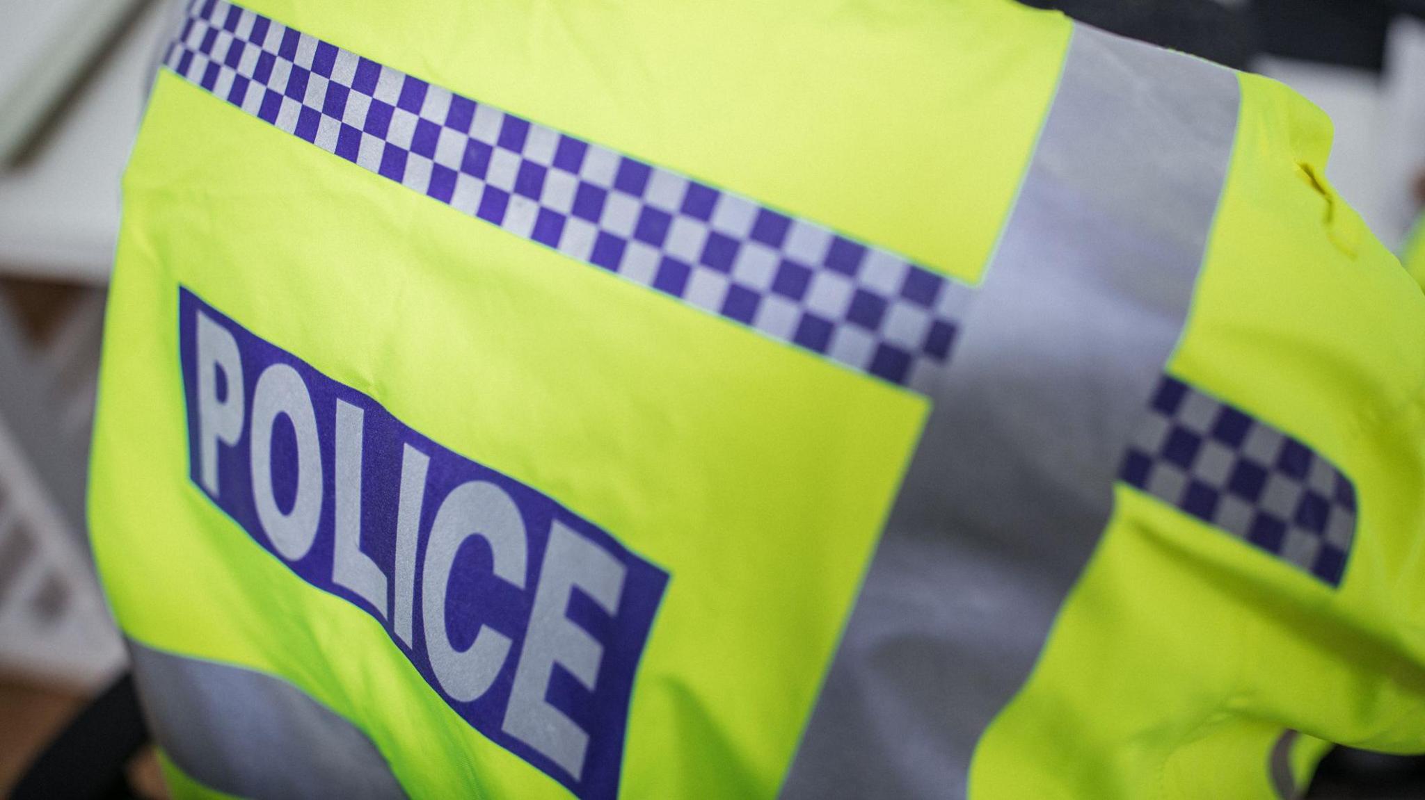 The back of a yellow high-visibility police vest, worn by and officer and taken from a downward angle. The vest has silver and blue checked stripes on it, and the word "police" written in capital letters across the back. 