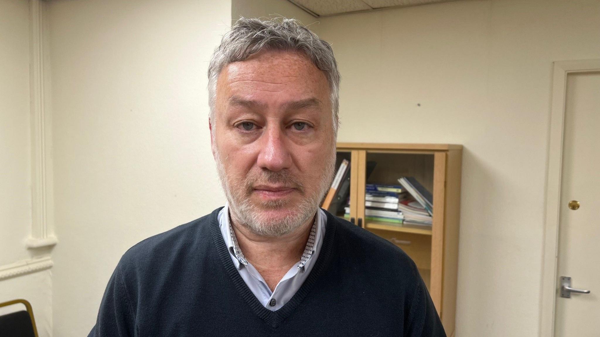 A man with short grey hair and a tightly cropped grey beard is looking directly ahead. He is wearing a blue V-neck jumper on top of a blue shirt with a collar. He is standing in a room with beige walls, with a bookcase visible in the background.