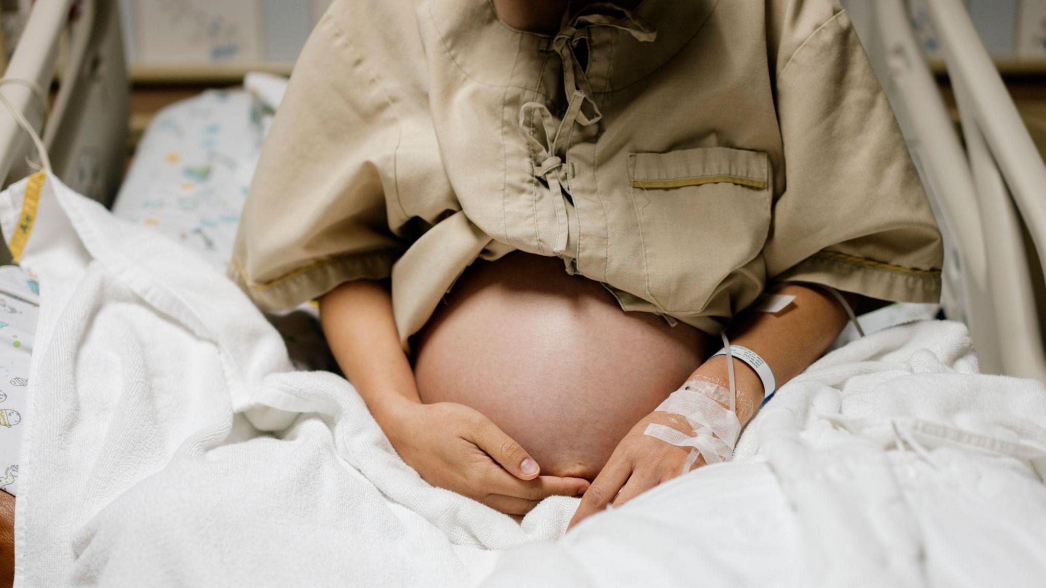 Close-up of belly a heavily pregnant person in a hospital bed, with an IV attached to her arm.