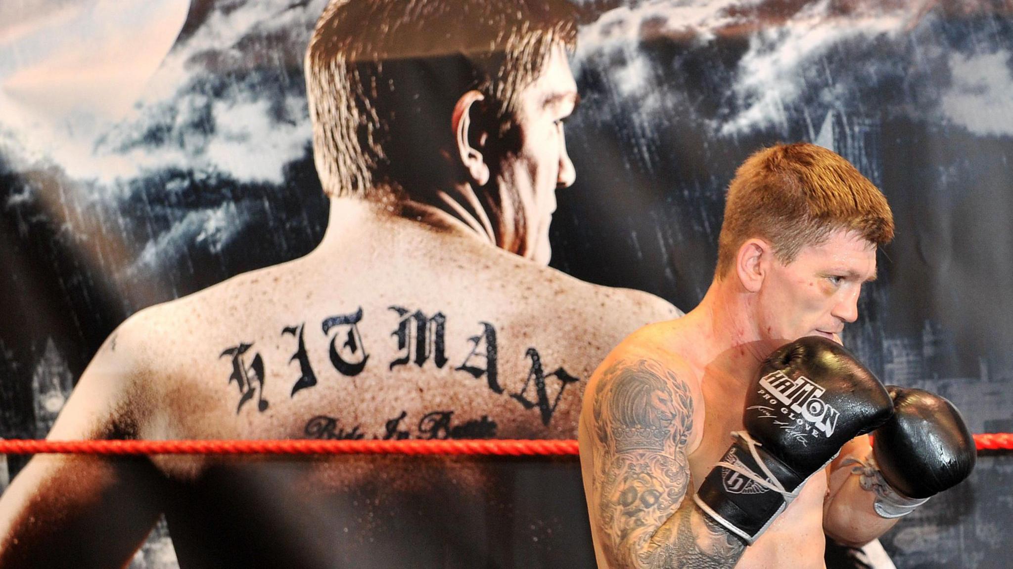 A wide shot of Ricky Hatton wearing black boxing gloves. In the background is an image of Hatton's back, with the tattoo 'hitman' written across it.