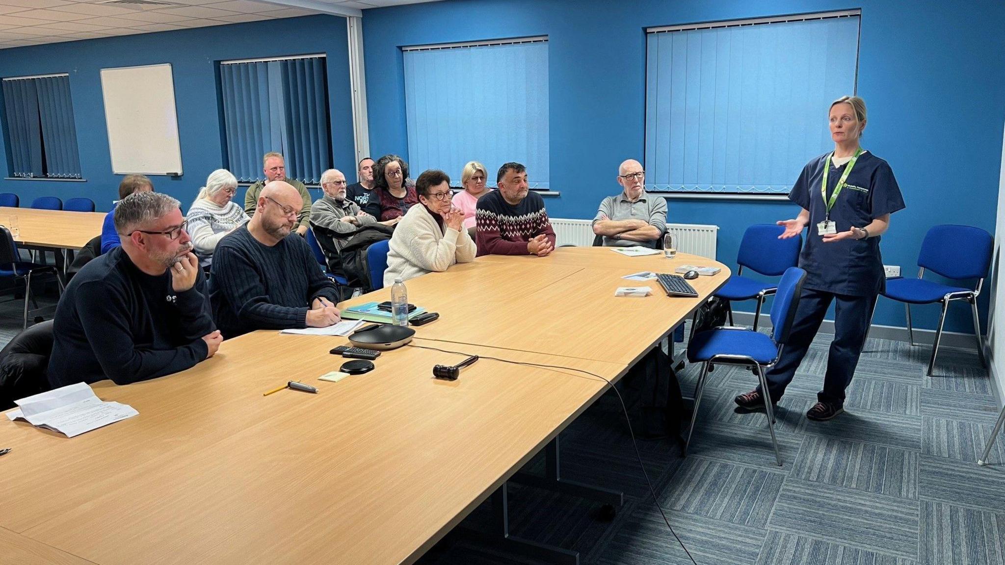 A female doctor, who has blonde hair and is wearing surgical scrubs, is stood in front of a class of patients who are there to learn about diabetes.  There are twelve people sitting behind the table listening.  Most of them are in their fifties and sixties.