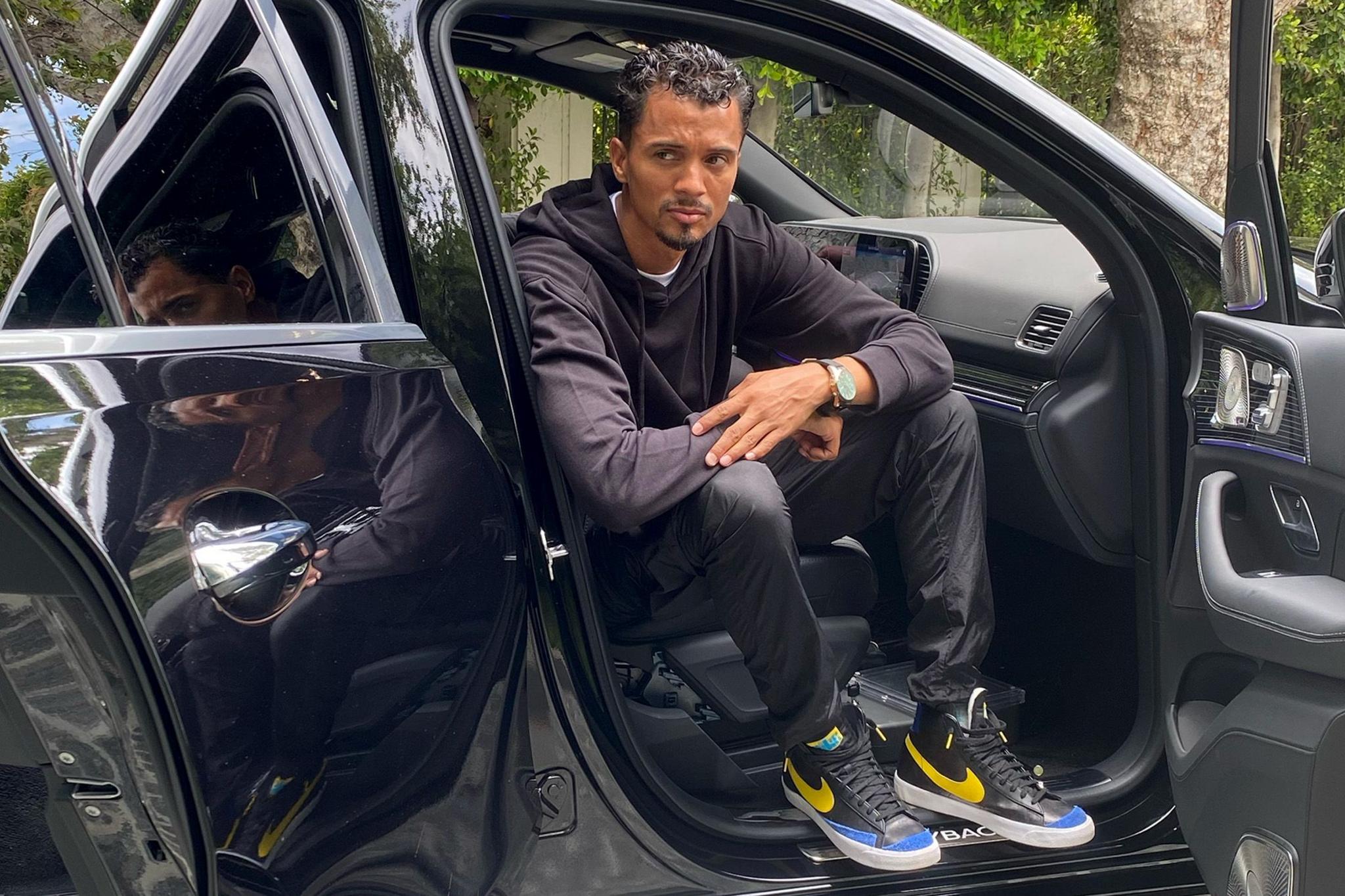 Phil Pines in a black hoodie, pants, and sneakers with yellow and blue accents sitting in the passenger seat of a black car with the door open.