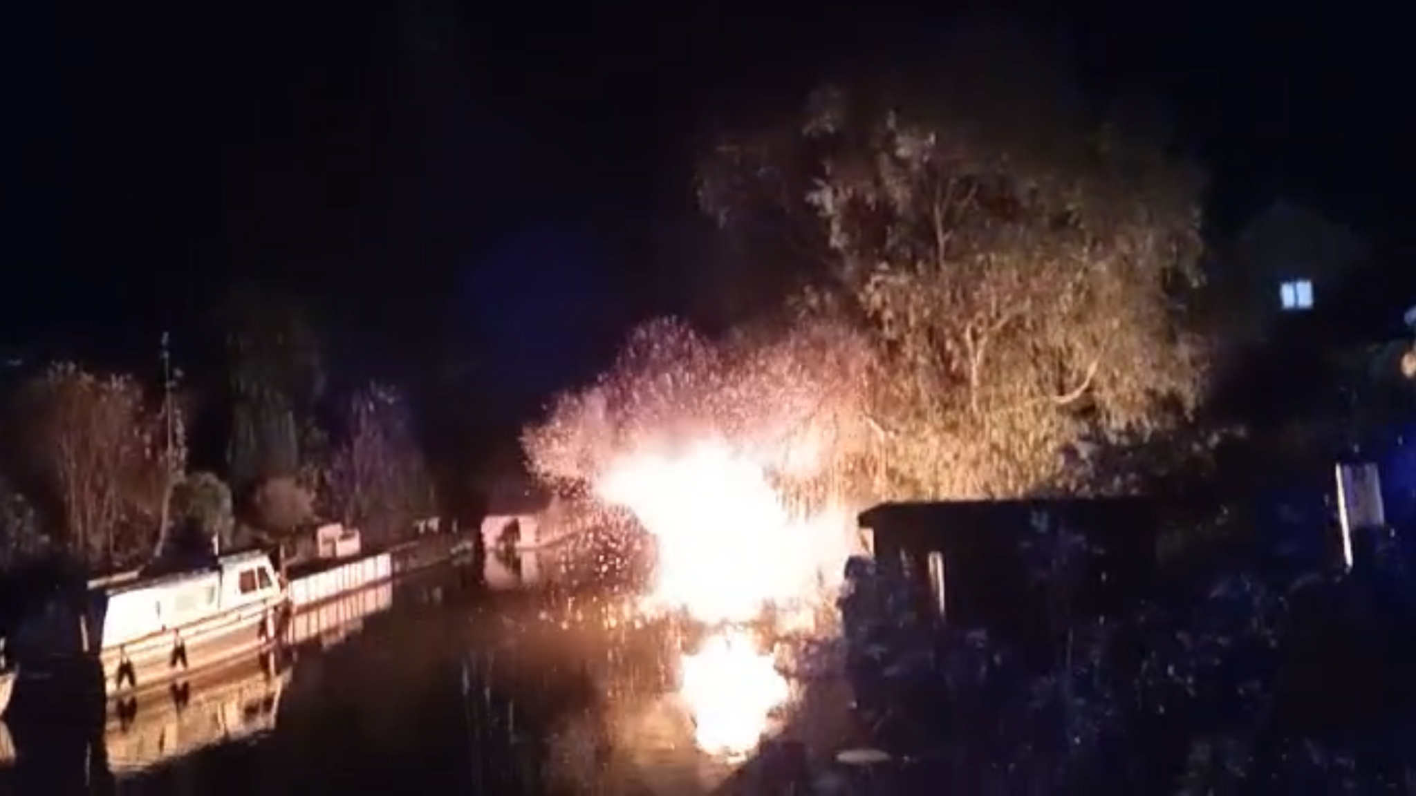 A large ball of flames on the river, illuminating a nearby tree, sparks can be seen shooting out of the flames, there are two other cruiser boats moored up near the blaze.