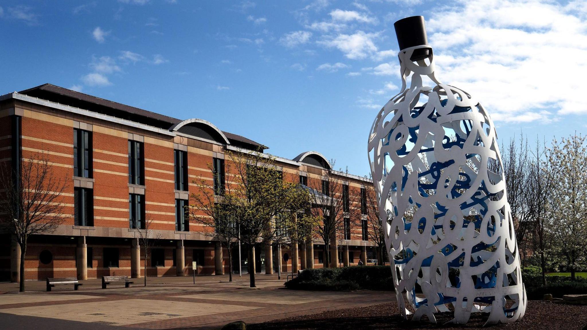 Teesside Crown Court. It is a multi-storey red brick building with a number of windows running along its front. A sculpture of a large bottle stands nearby.