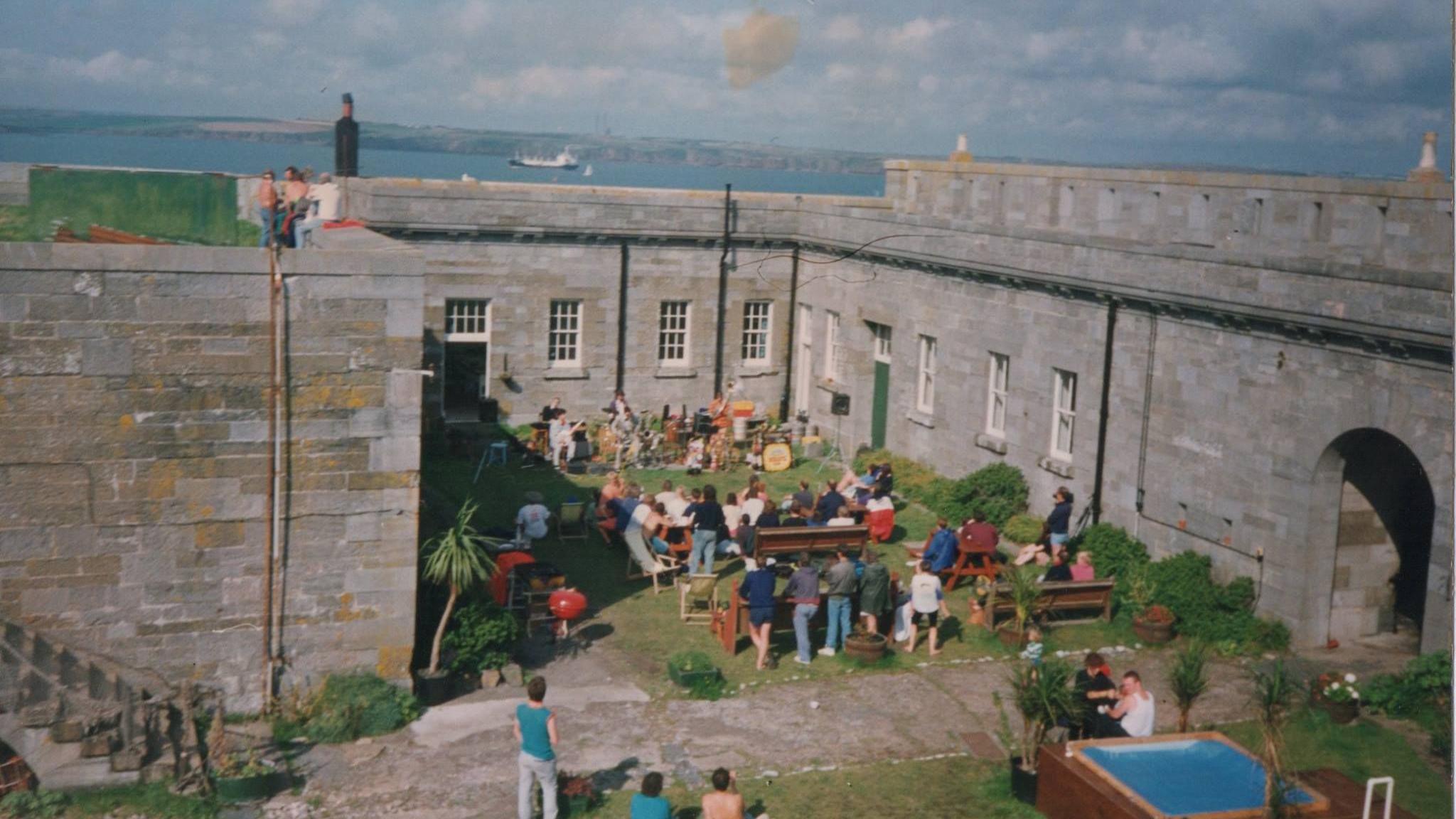 A group of people sit on benches in a courtyard, surrounded by large grey fortress walls. They are facing a band set up along the far wall. A small square plunge pool can be seen in the right hand corner. The  sea can be seen in the distance. 