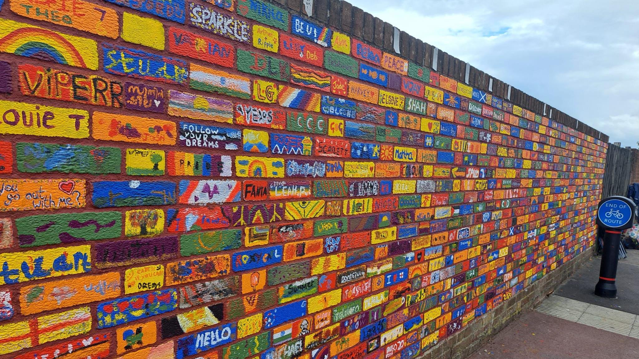A colourful brick wall. Each brick is painted with pictures and messages in an array of colours - mostly yellows, blues, greens and reds.