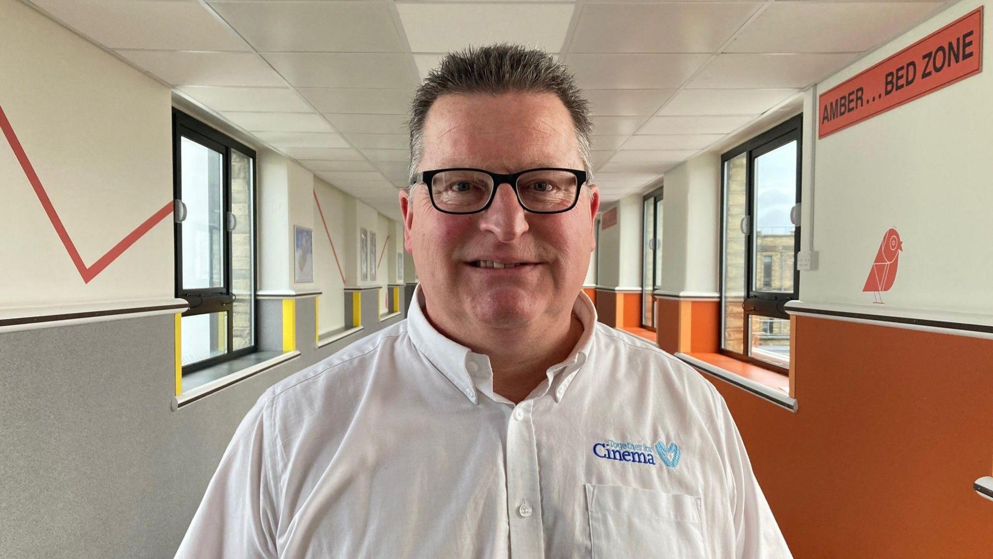 A man with short spikey hair and glasses wearing a white shirt with the words Together for Cinema stitched above the breast pocket. He is standing in a colourful hospital corridor.