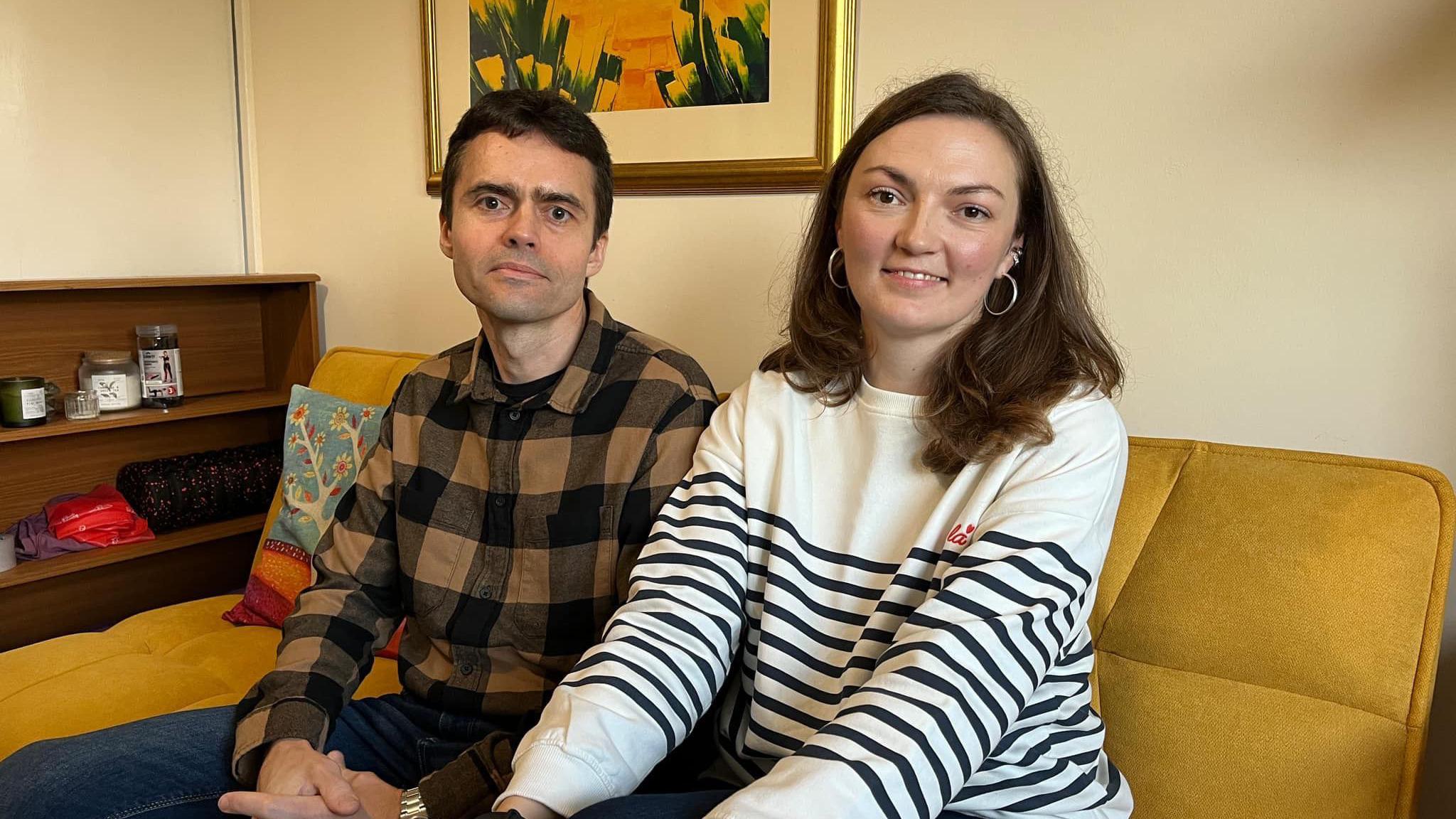 Ukrainian couple sitting together on a yellow sofa in a house in Stroud. The man has a brown shirt on, while the woman has a white top with black strips on it. Behind the couple there is a painting on the wall. 