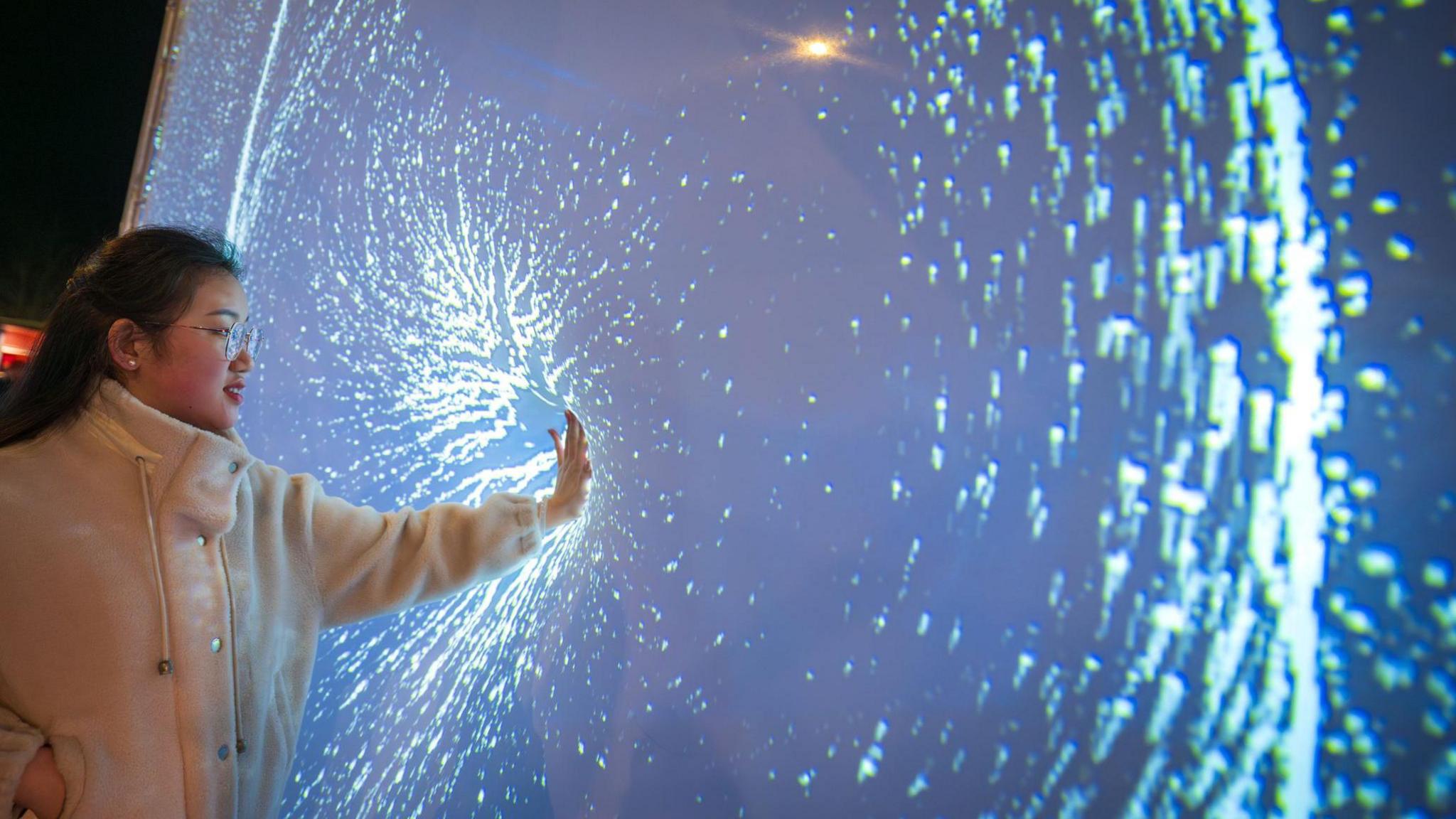 A woman is touching a light installation. White lights are near the spot where her hand touches the screen