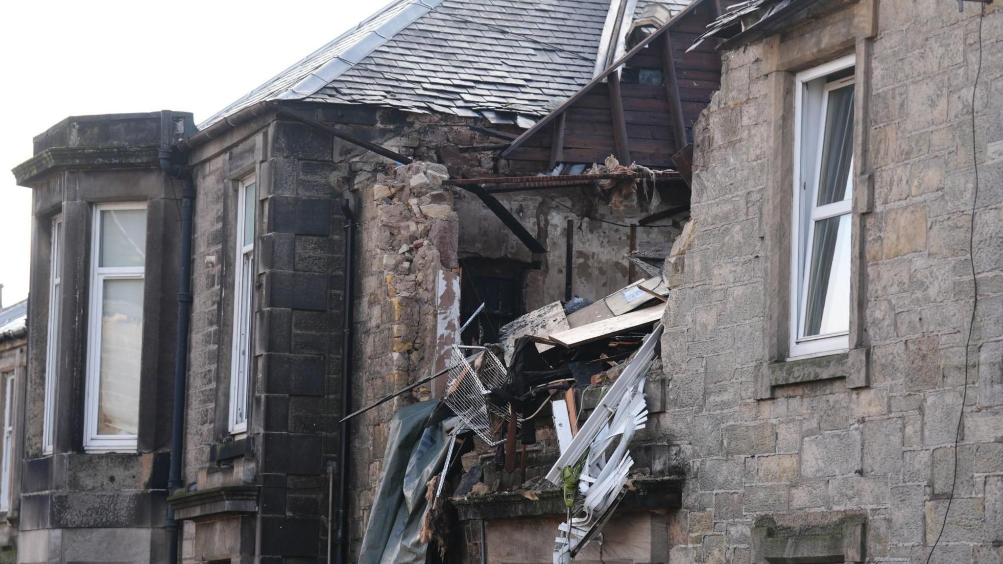 A grey sandstone building with a large hole on the upper floor and the roof. Debris from inside the flat can be seen sticking out of the gap.