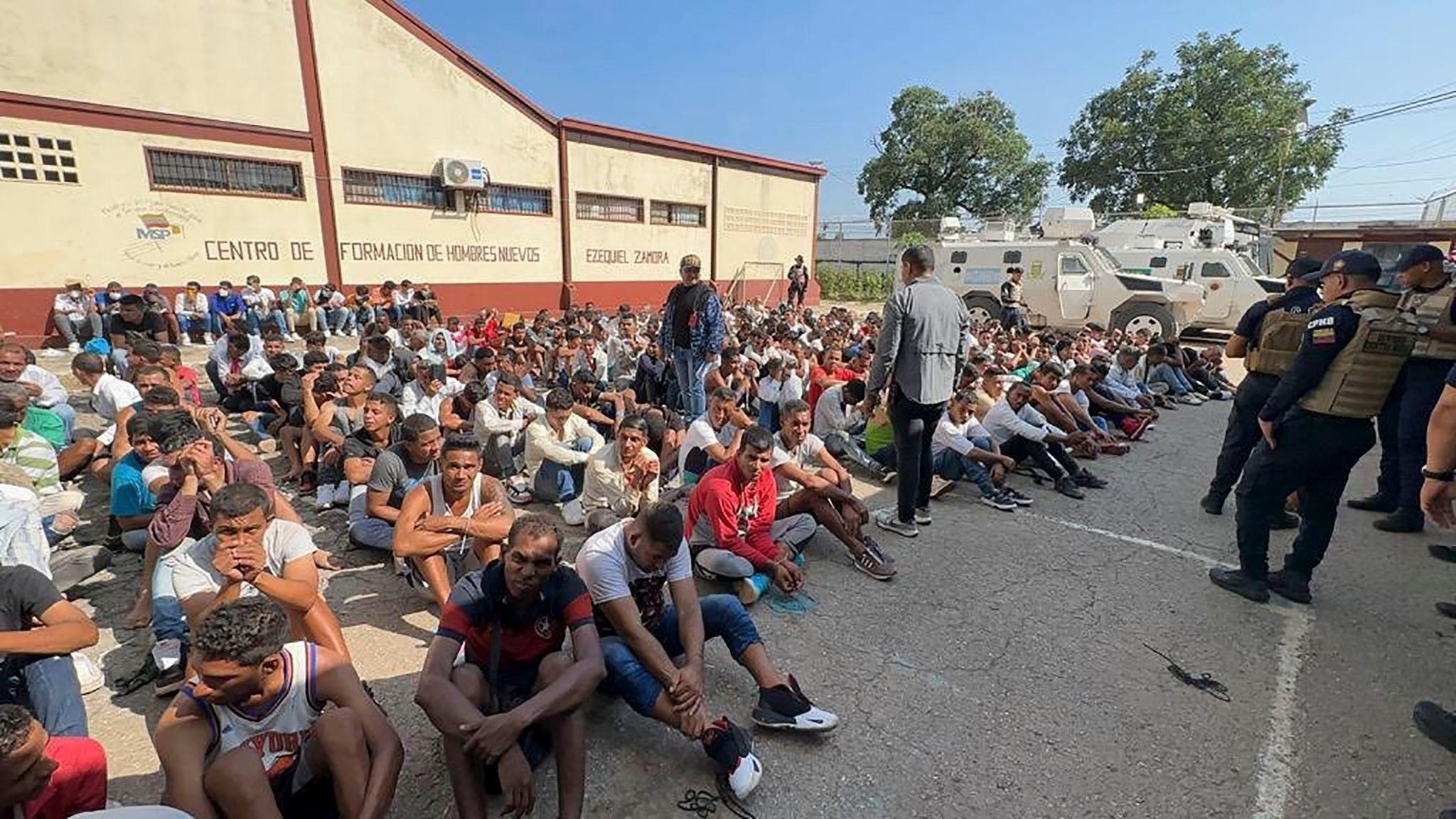 Men sitting on black pavement in lines with guards in bullet proof bests standing in front of them 