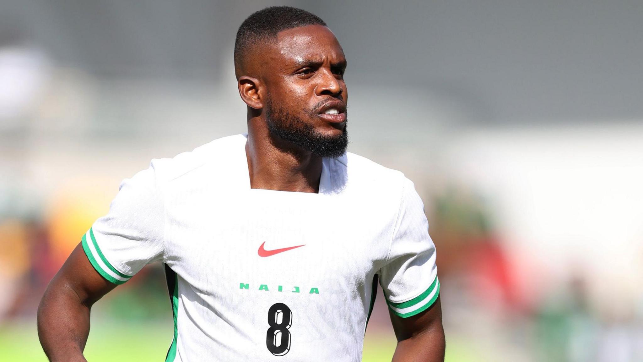 Frank Onyeka is seen from the waist up wearing a white Nigeria shirt with green trim and the number 8 on the chest beneath the word Naija printed in green capital letters