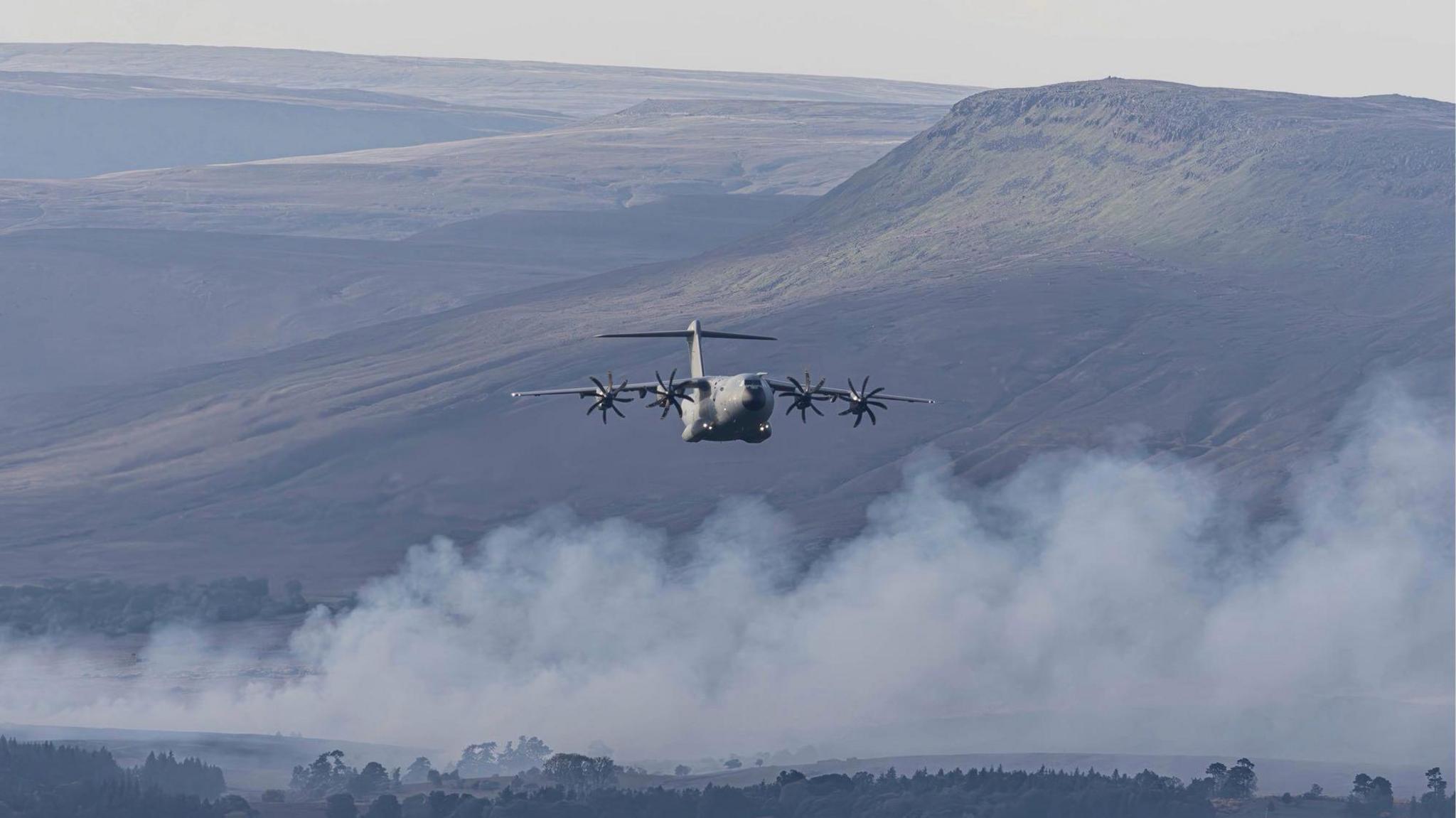 An M405 - A400M plane passing in front of hills and smoke coming from the forest below.