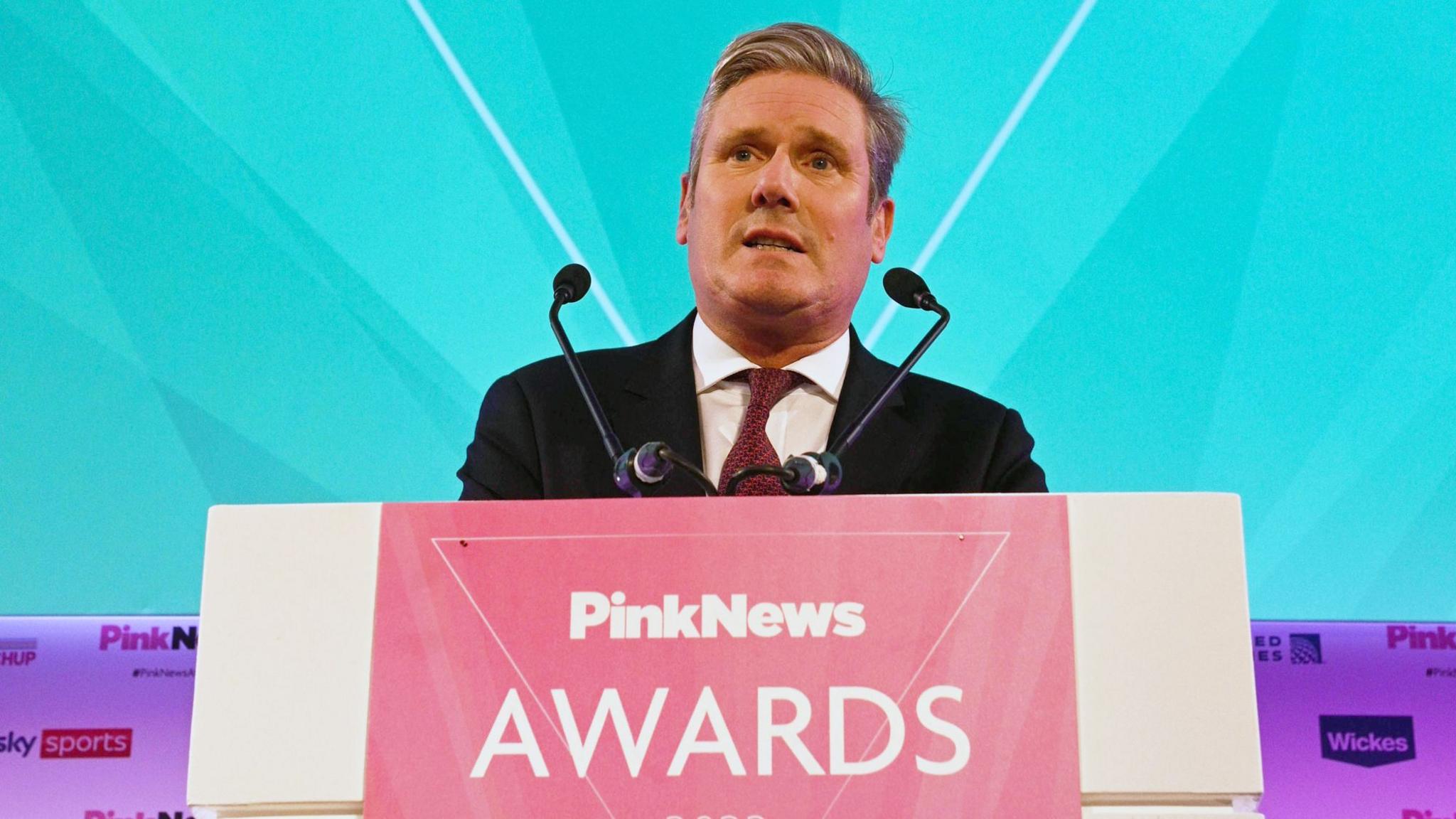 Sir Keir Starmer standing at a lectern with a sign reading "PinkNews Awards", wearing a  dark suit and tie, with a turquoise background behind him. 