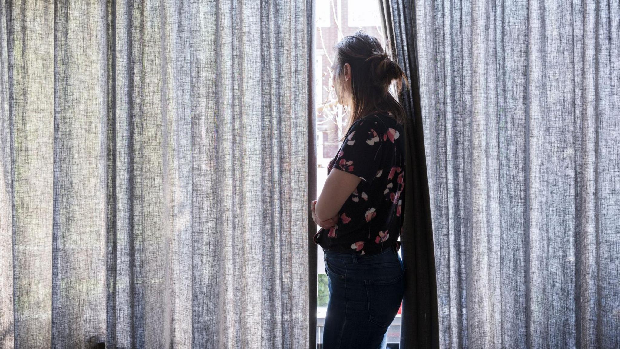 A woman looking out a window in between partially drawn curtains with her arms folded.