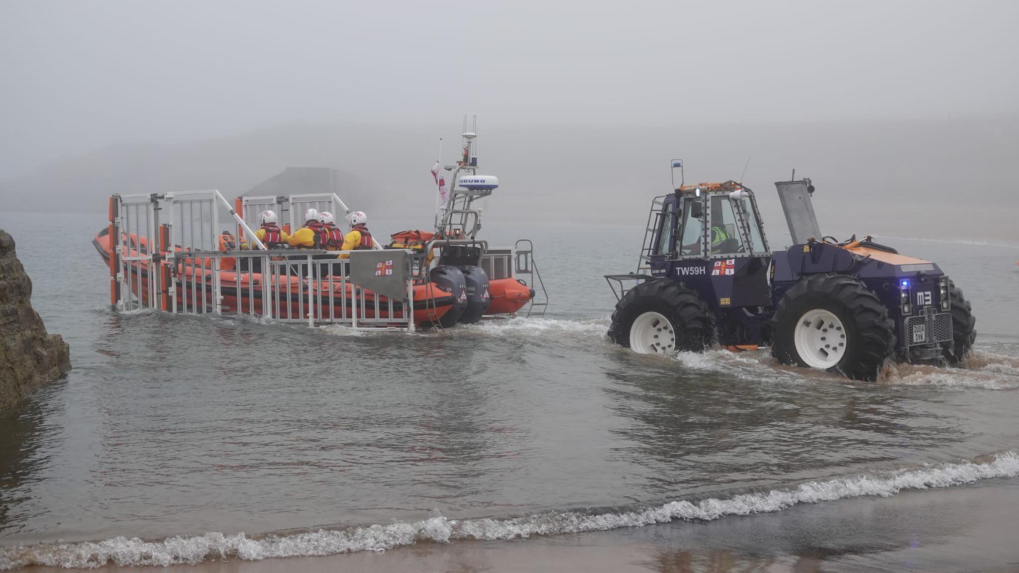 The inflatable lifeboat being launched into the water during foggy weather. It is being transported from the beach on a metal trailer attached to a blue tractor.