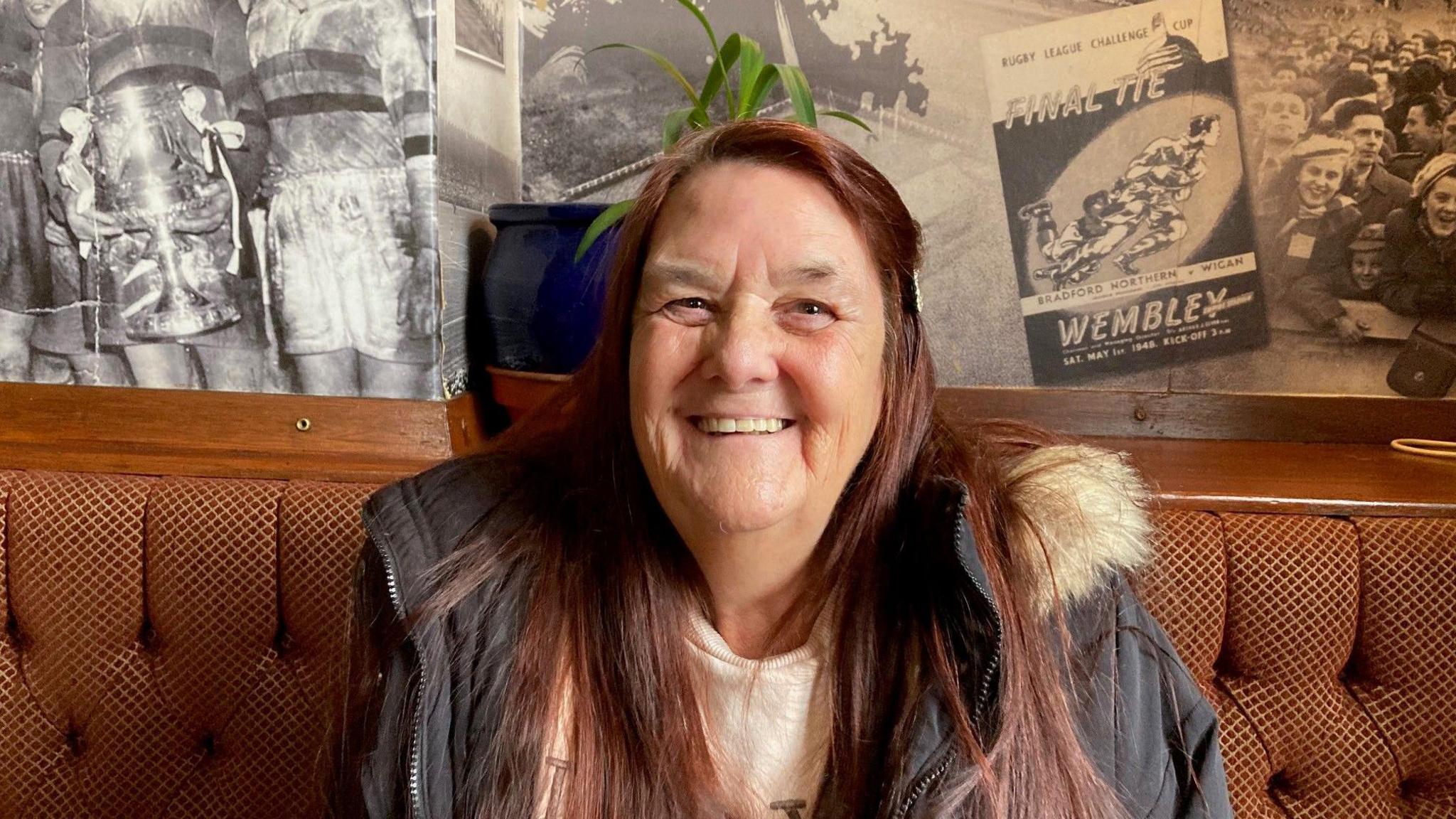 A smiling woman with long, brown hair on some brown banquette seating.